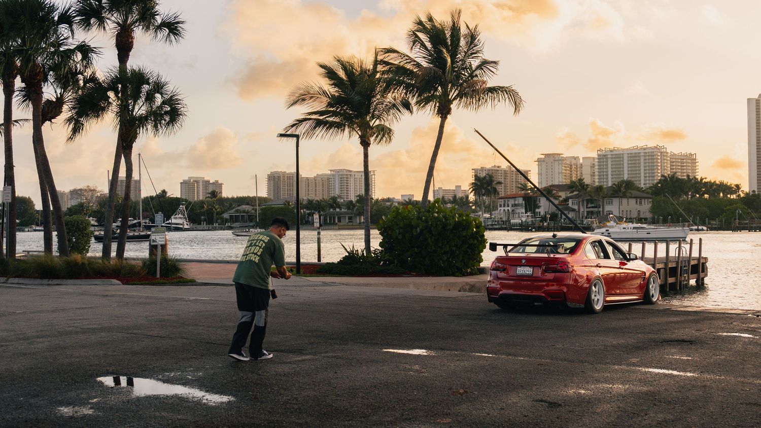 A person skateboards near a red sports car parked by a waterfront with palm trees and city skyline at sunset.