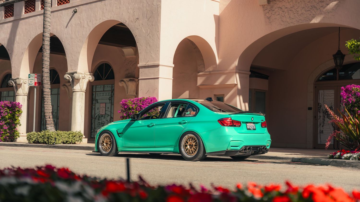 Teal BMW M3 sedan with gold wheels parked outside a luxury building with arches and tropical flowers.