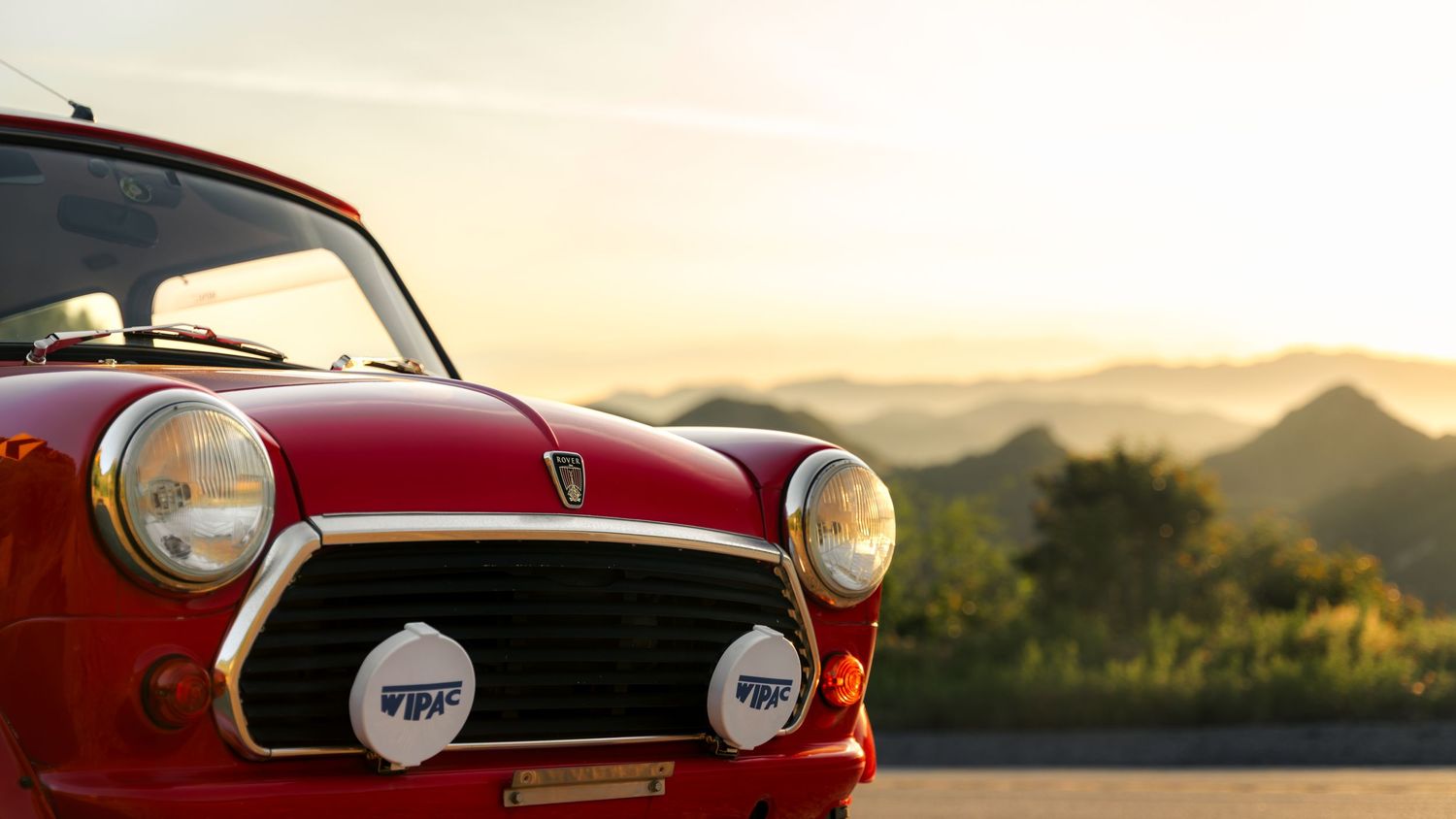 A vintage red MGB sports car parked in front of a scenic mountain landscape at sunset.