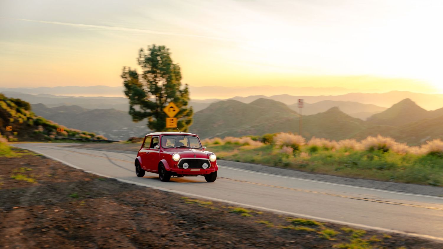 A red vintage sports car drives along a winding mountain road at sunset with scenic mountain vistas in the background.