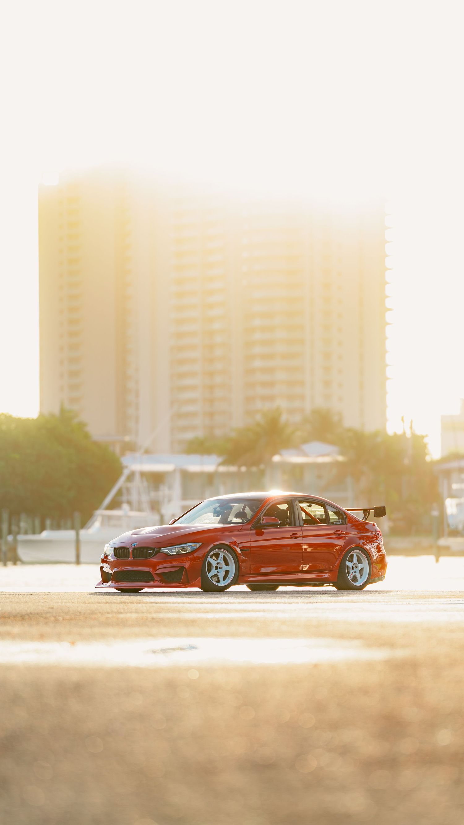 Red BMW M3 sports car with wide body kit parked in golden sunlight with blurred city buildings in background.