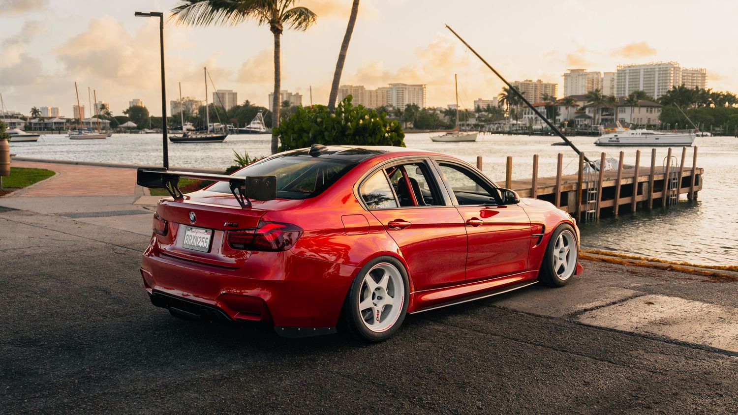 Red BMW M3 sedan with white wheels parked near a waterfront dock at sunset in a coastal city.
