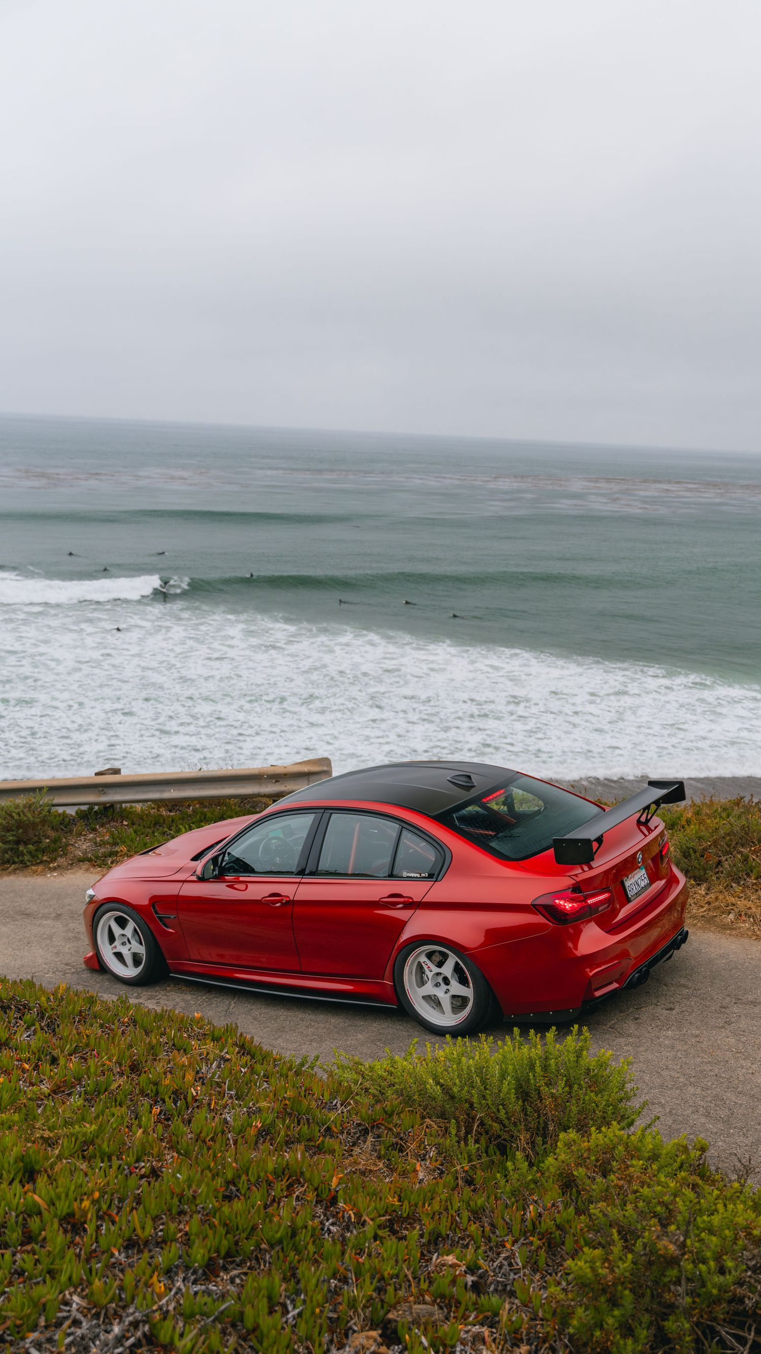 Red BMW M3 with carbon spoiler parked on coastal overlook with ocean waves behind.