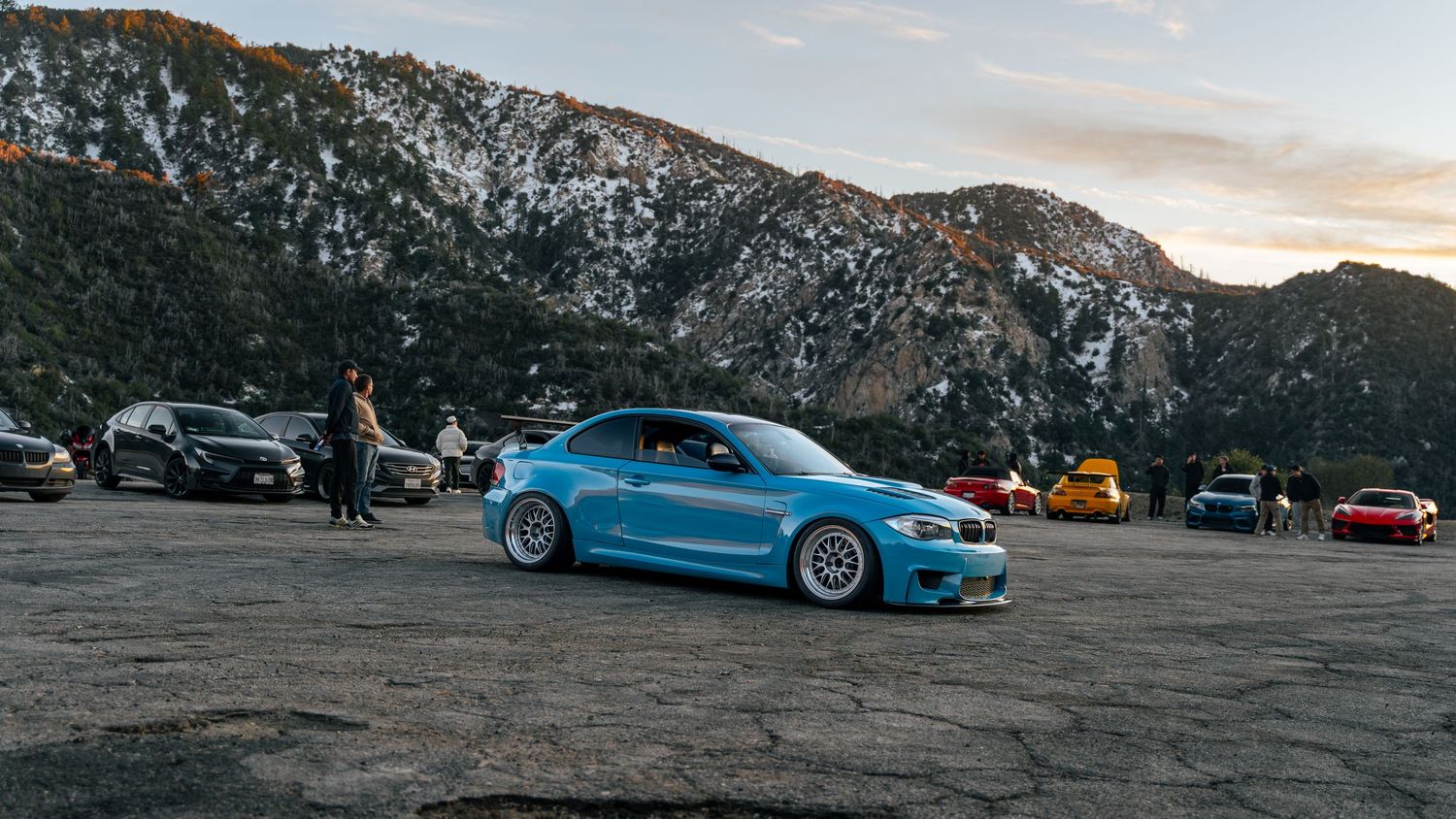 Sports cars lined up on a mountain road at sunset with rocky cliffs in the background.