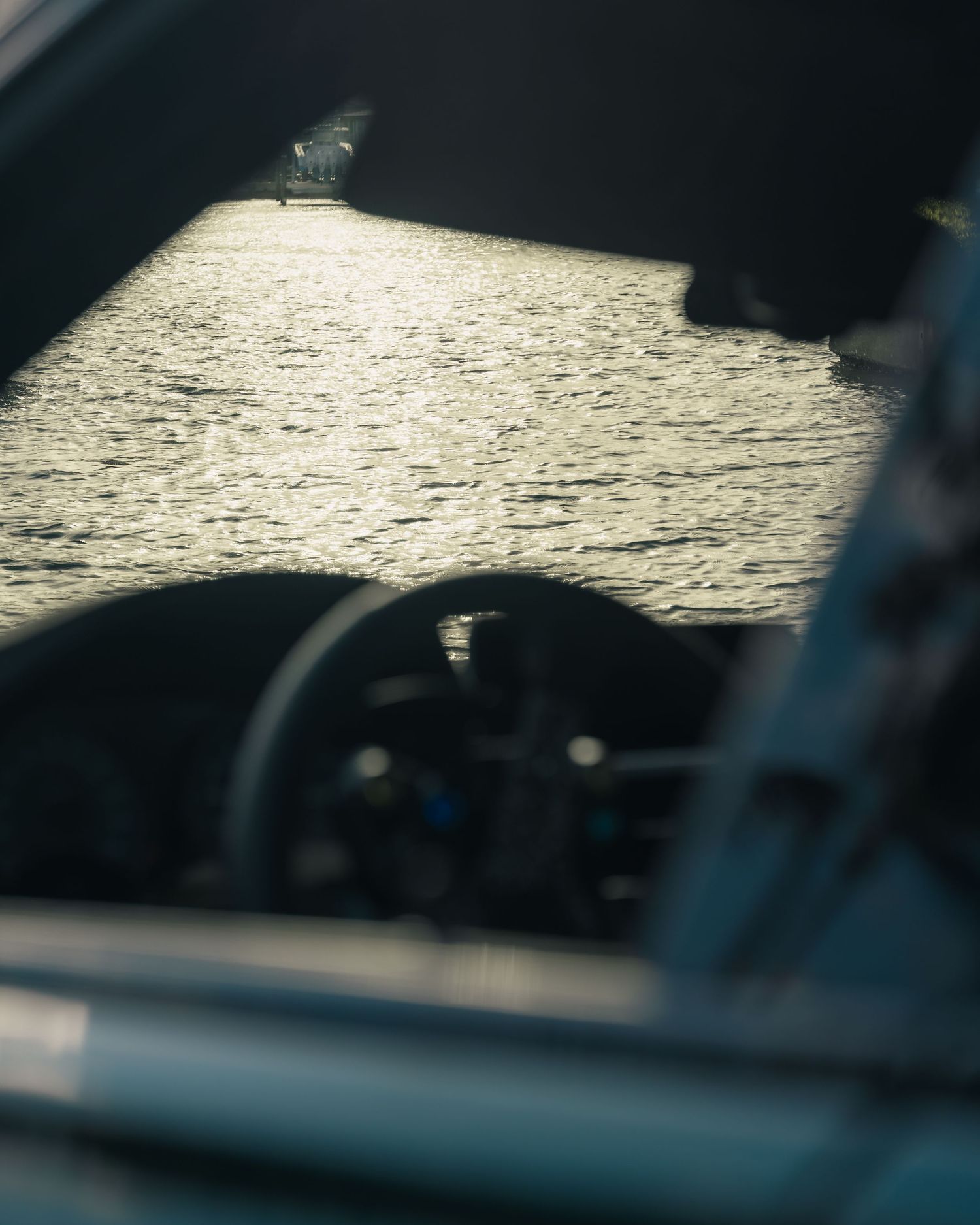 View through a car window of sunlight reflecting off rippling ocean water, with a steering wheel visible in the foreground.