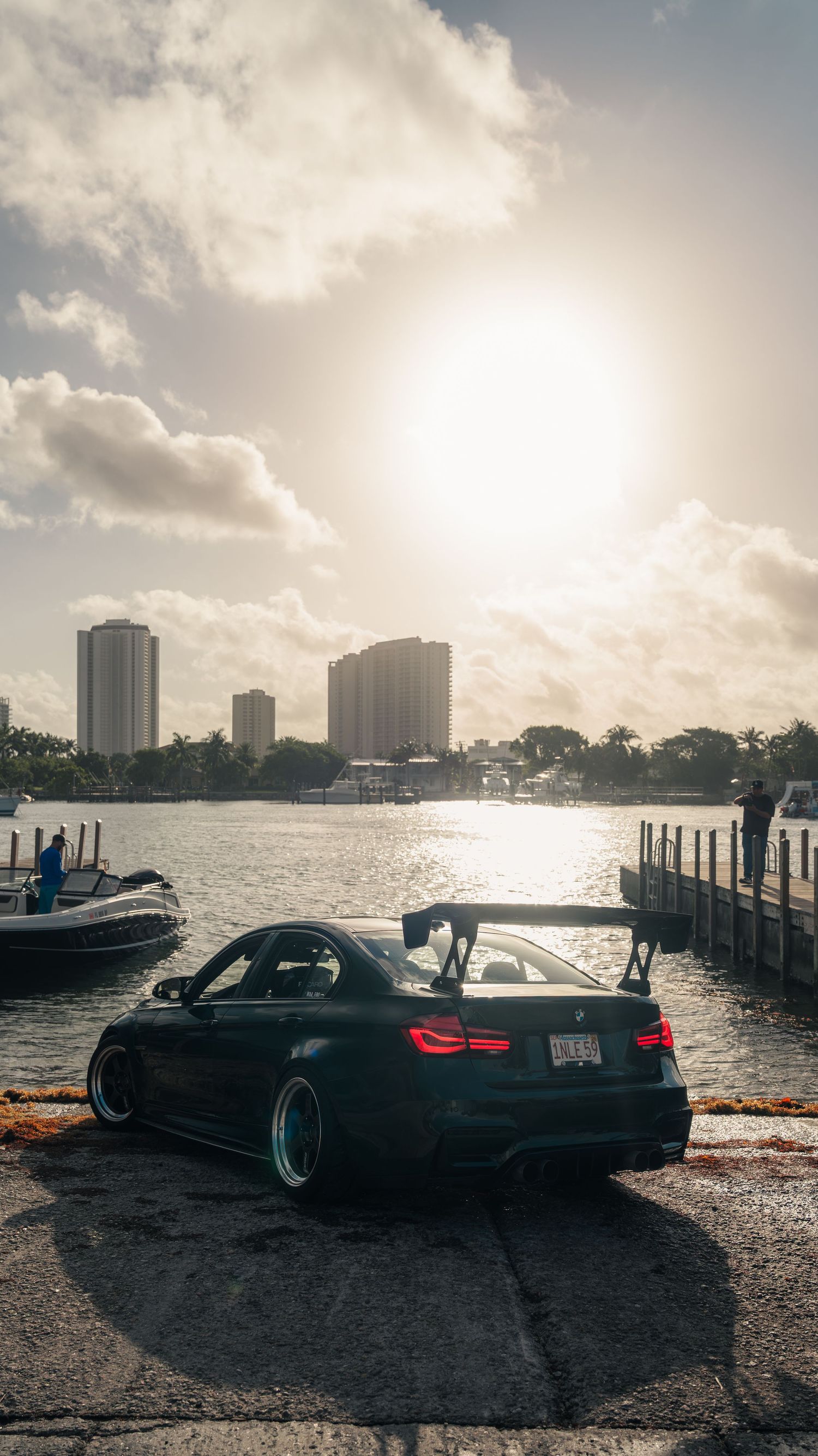 Modified BMW M4 with large rear spoiler parked at waterfront dock, city skyline and dramatic sunlight reflecting on water.