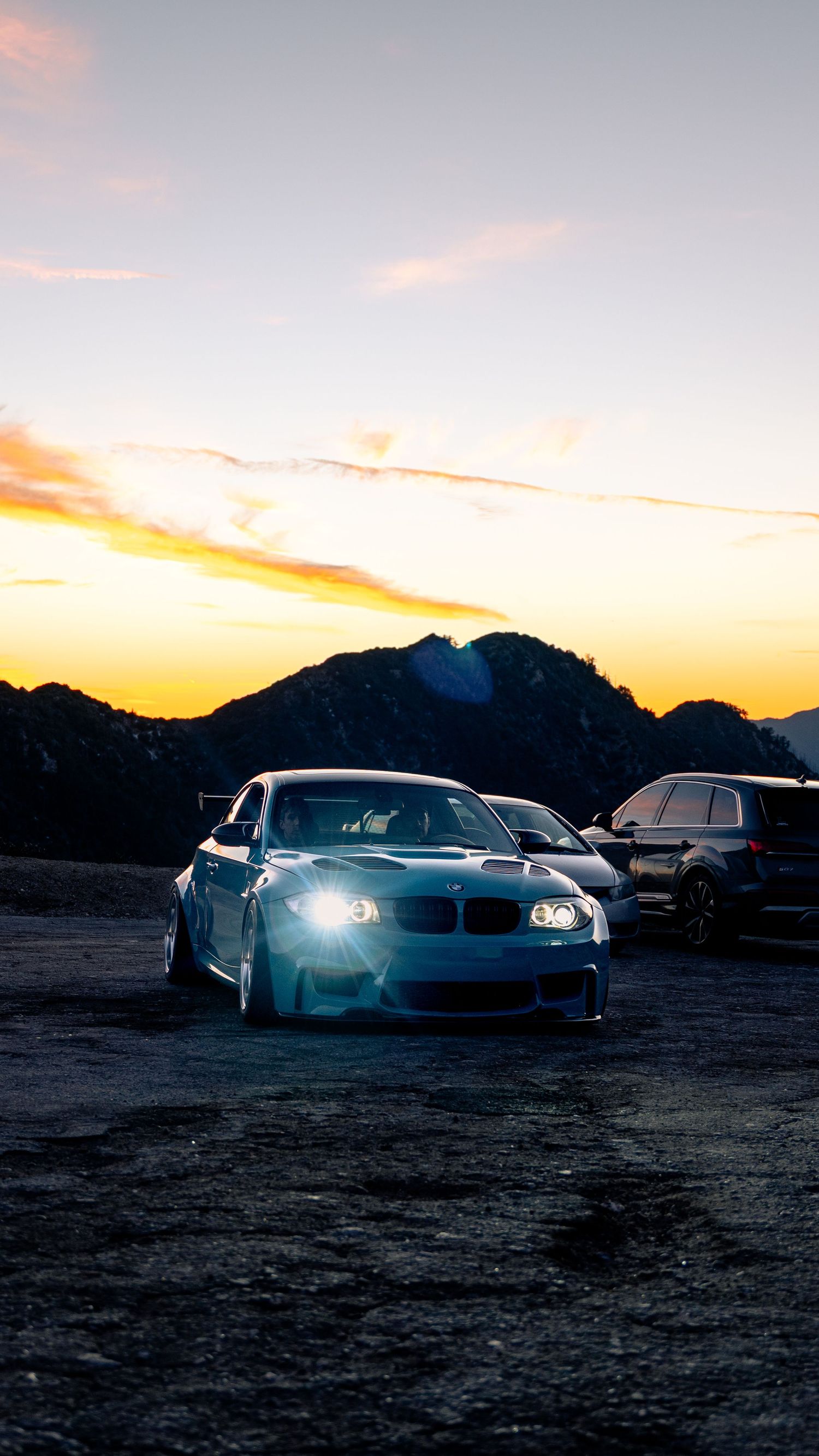 BMW sports cars parked against a dramatic mountain sunset with glowing headlights.