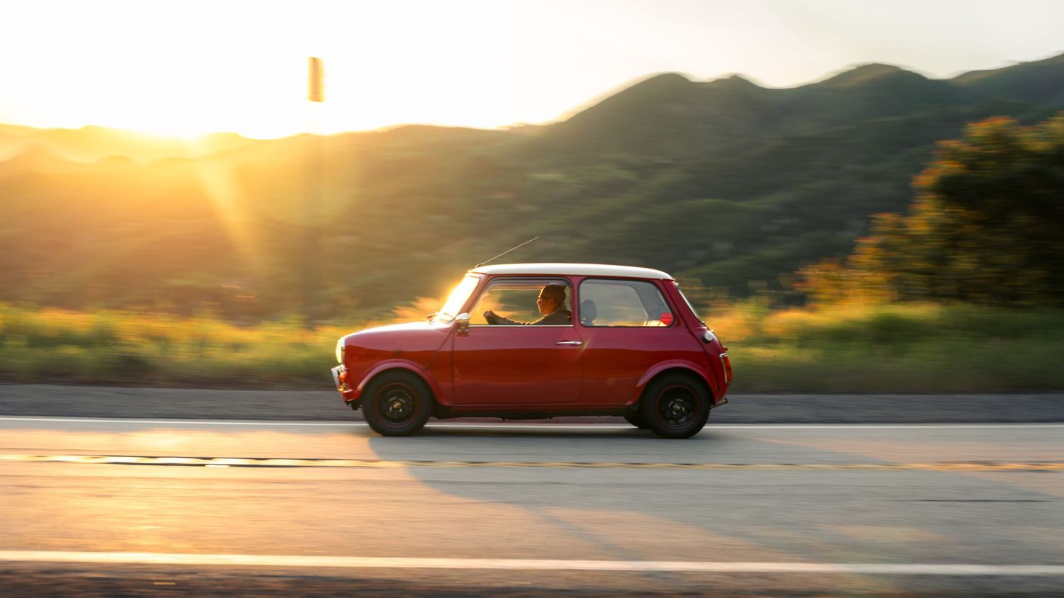 A red vintage car drives along a scenic mountain road at sunset with golden light illuminating the landscape.