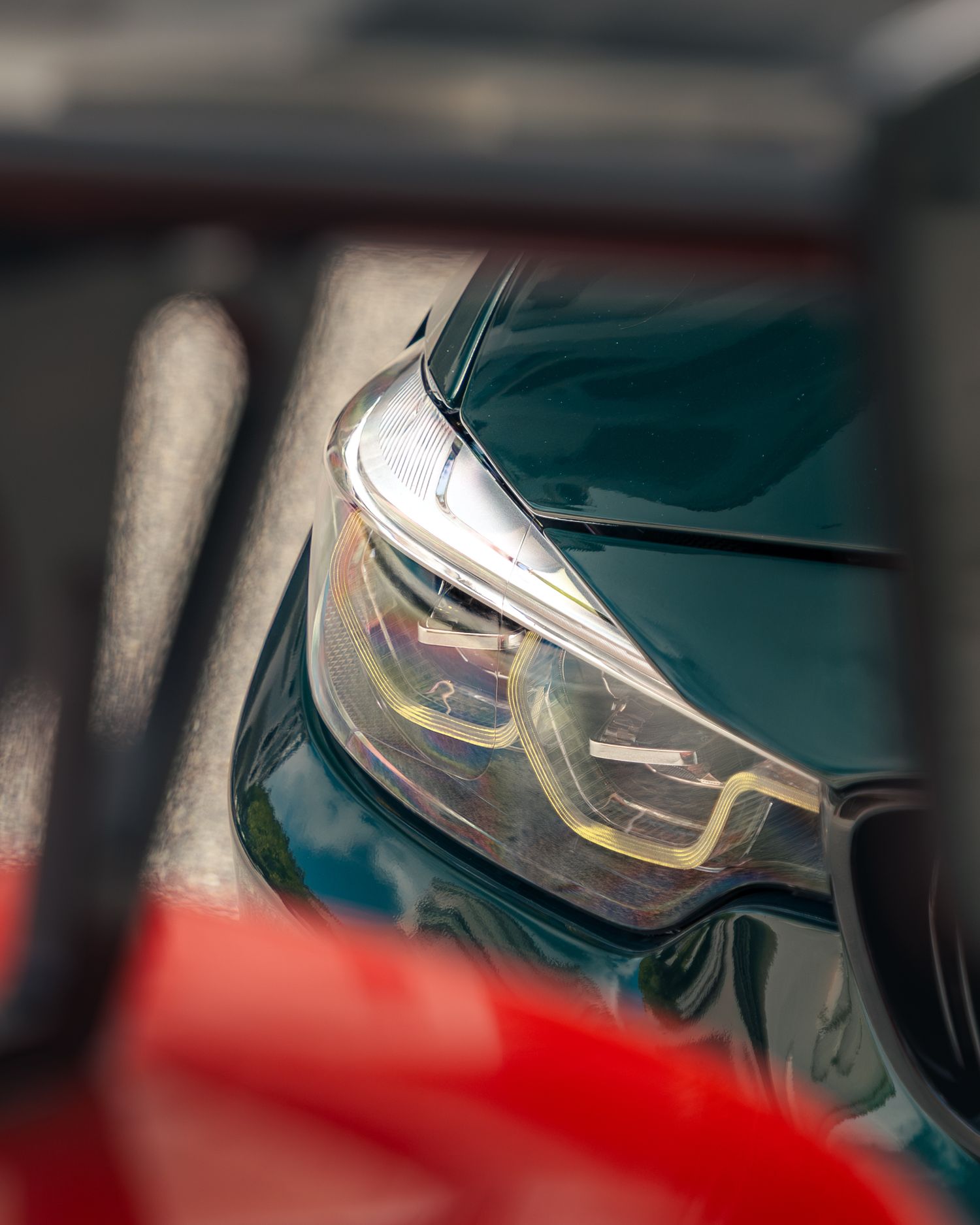 Close-up of a teal BMW headlight with angular LED daytime running lights, partially framed by red car bodywork.