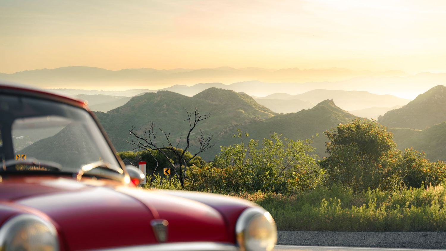 A panoramic view of rolling hills and mountains at sunset with a vintage red car in the foreground.