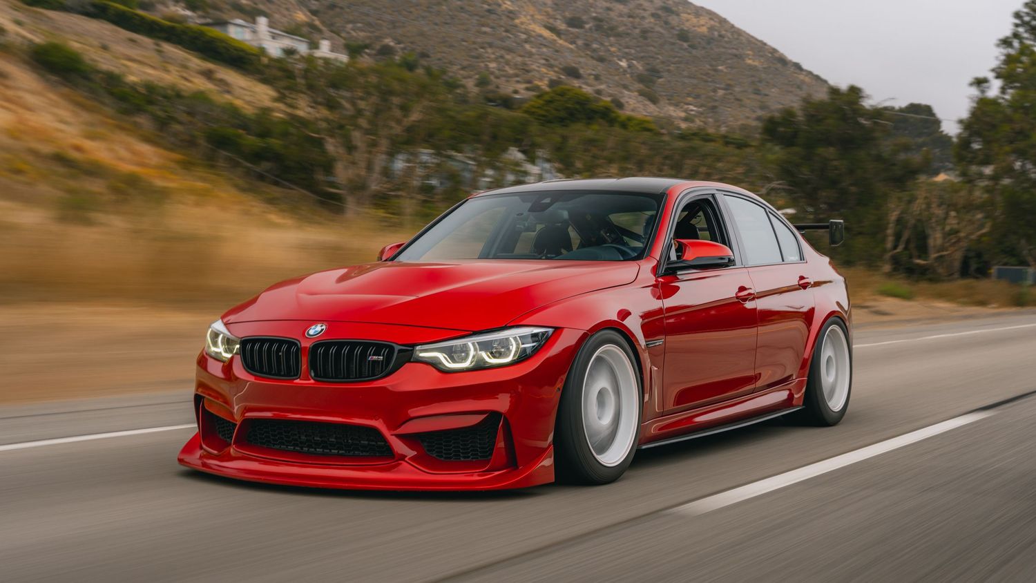 Red BMW M3 sedan with wide body kit driving on a mountain road, showcasing aggressive styling and white wheels.