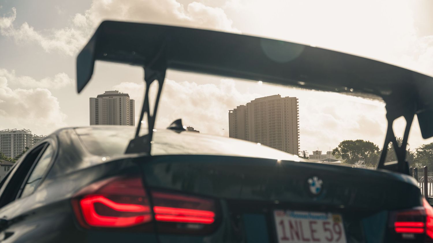 Close-up rear view of a BMW M car with large spoiler and red taillights against a sunny urban skyline.