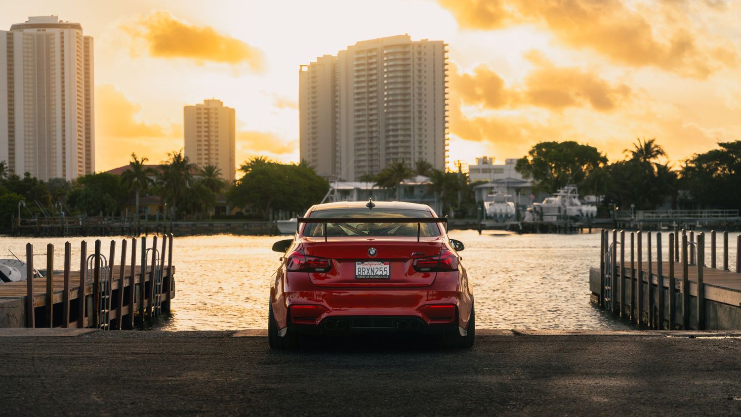 Red BMW sedan parked at waterfront dock with golden sunset sky and city skyline in Miami, Florida.
