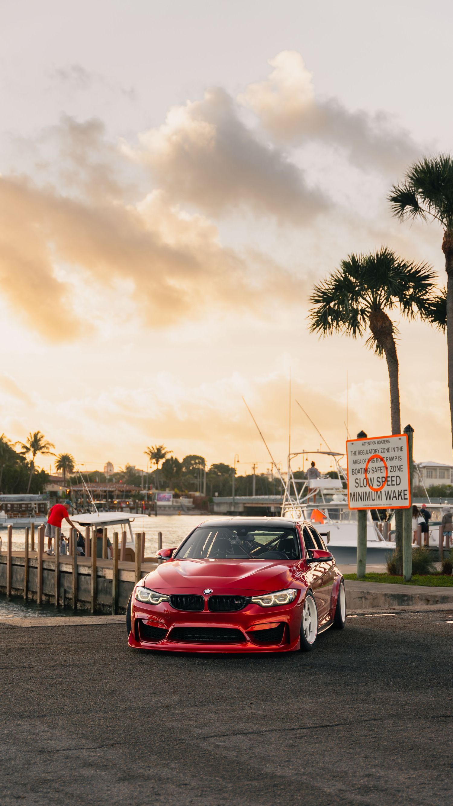 Red BMW M3 parked near a marina at sunset with palm trees and a speed limit sign in the background.