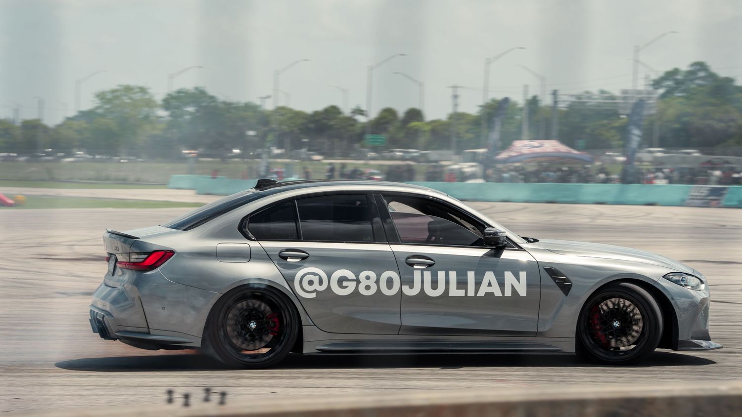 Silver BMW M3 drifting on a racetrack, leaving tire smoke trails, with @G80JULIAN watermark visible.