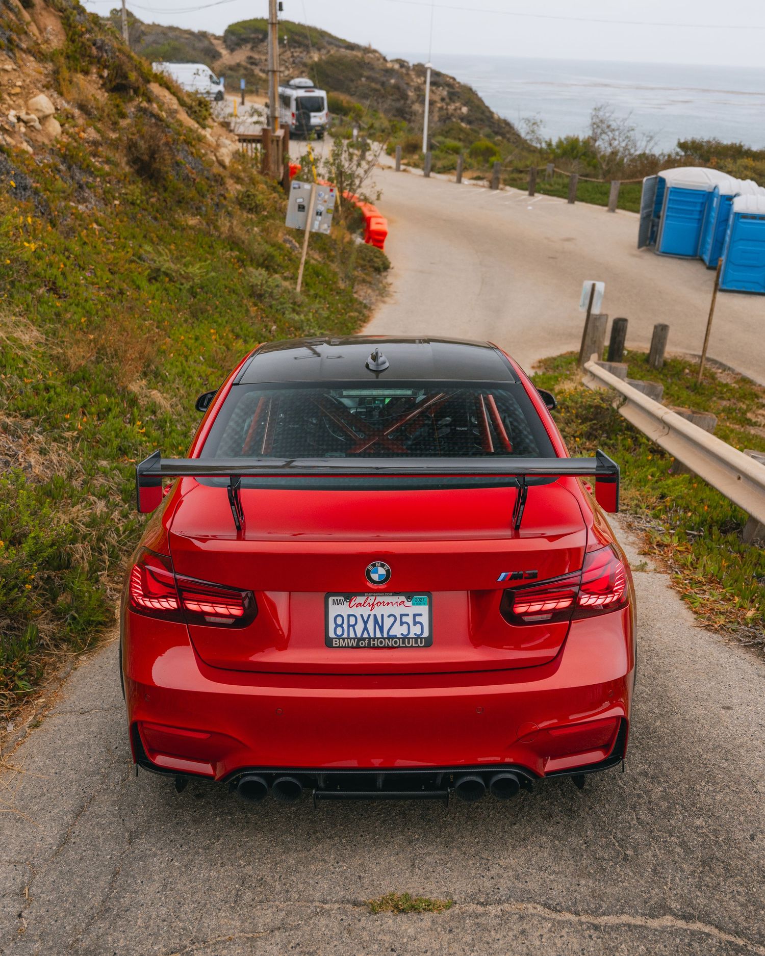 Red BMW M3 with large rear spoiler and diffuser parked on a coastal road overlooking the ocean in California.