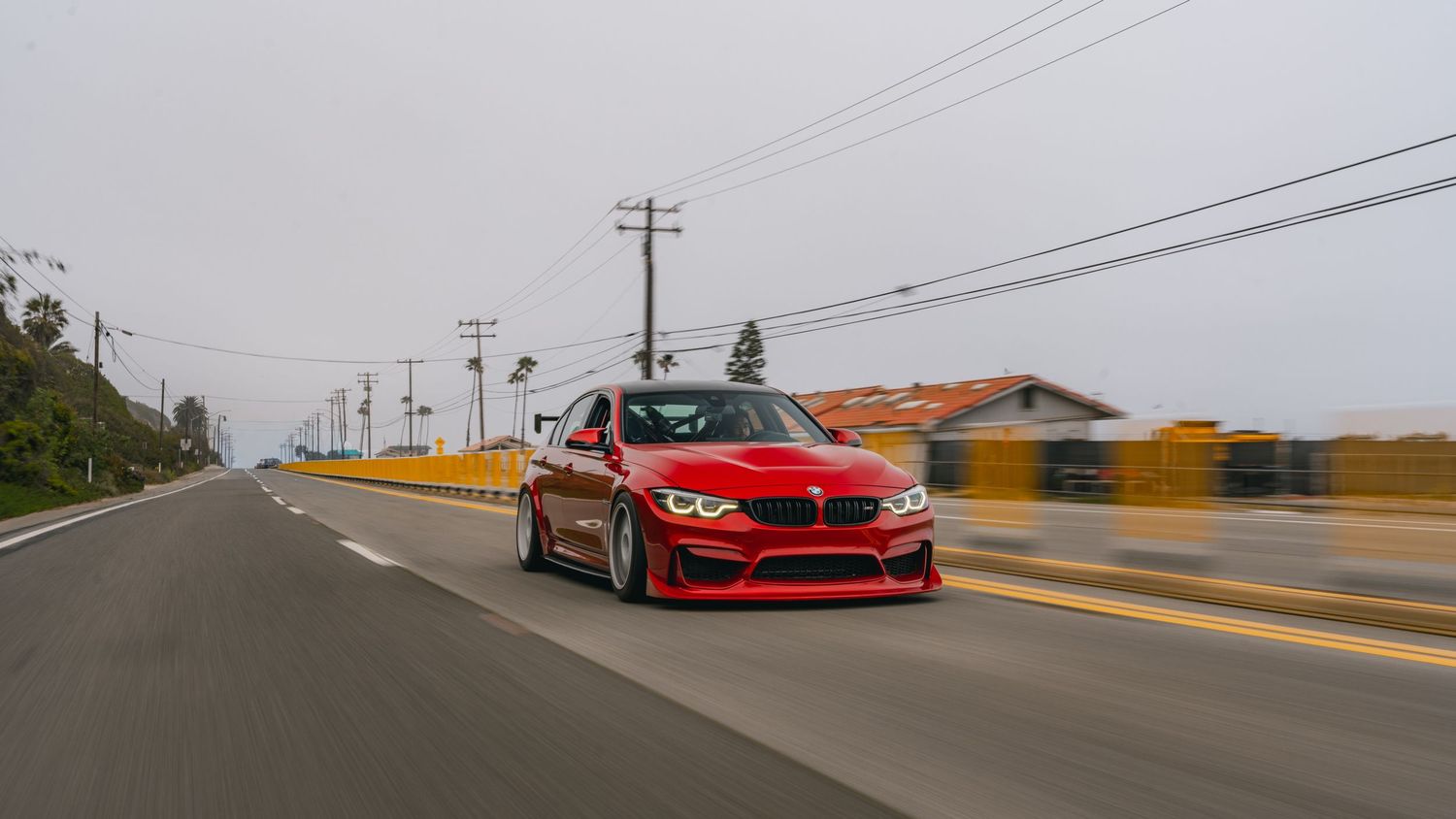 Red BMW M3 sedan speeding down a coastal highway under overcast skies with utility poles and buildings in background.