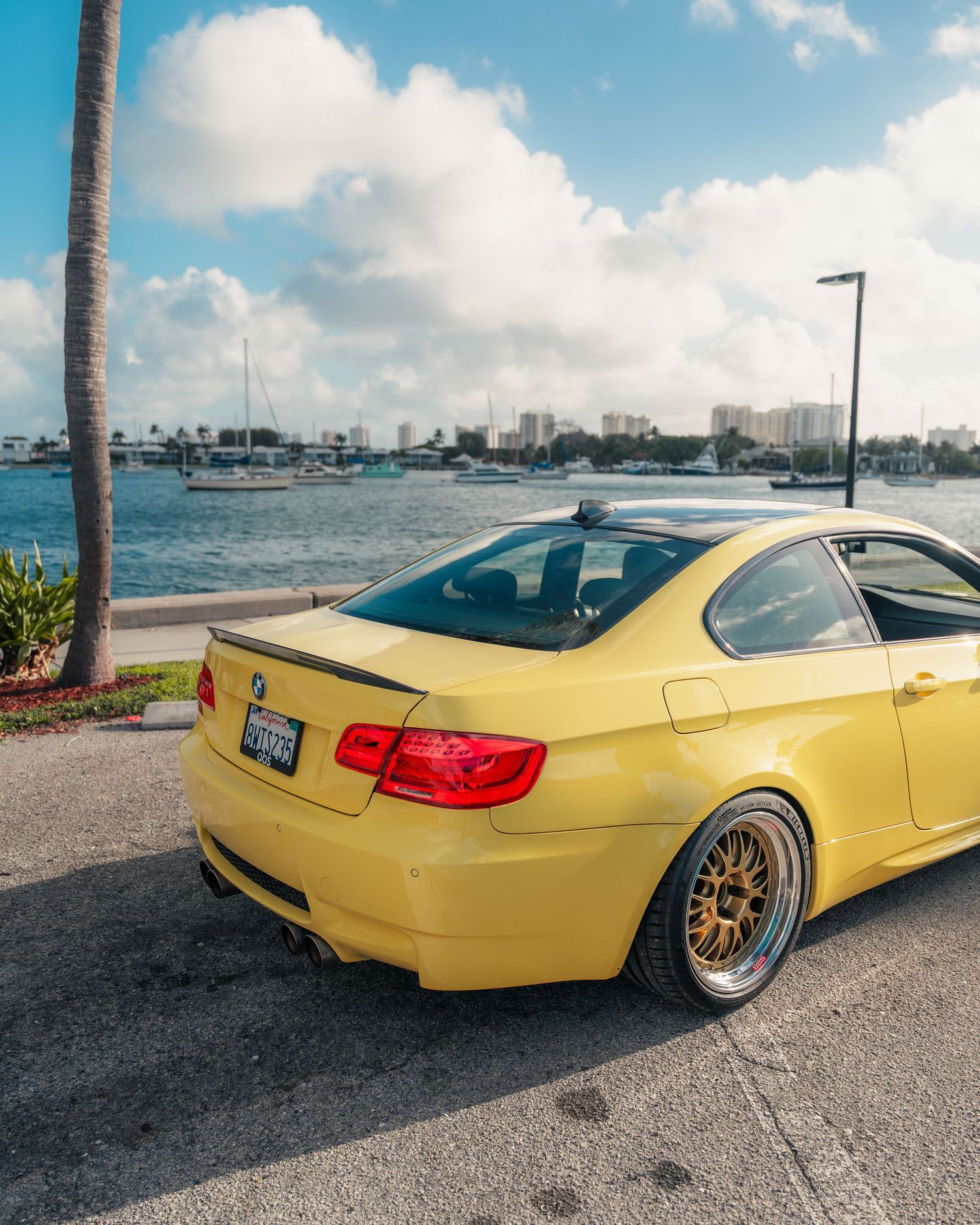 Yellow BMW coupe with bronze wheels parked near a waterfront on a sunny day with clouds and palm trees.