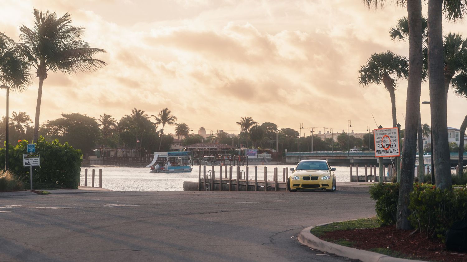 Yellow car parked near a waterfront marina with palm trees and dramatic cloudy sunset sky in Florida.