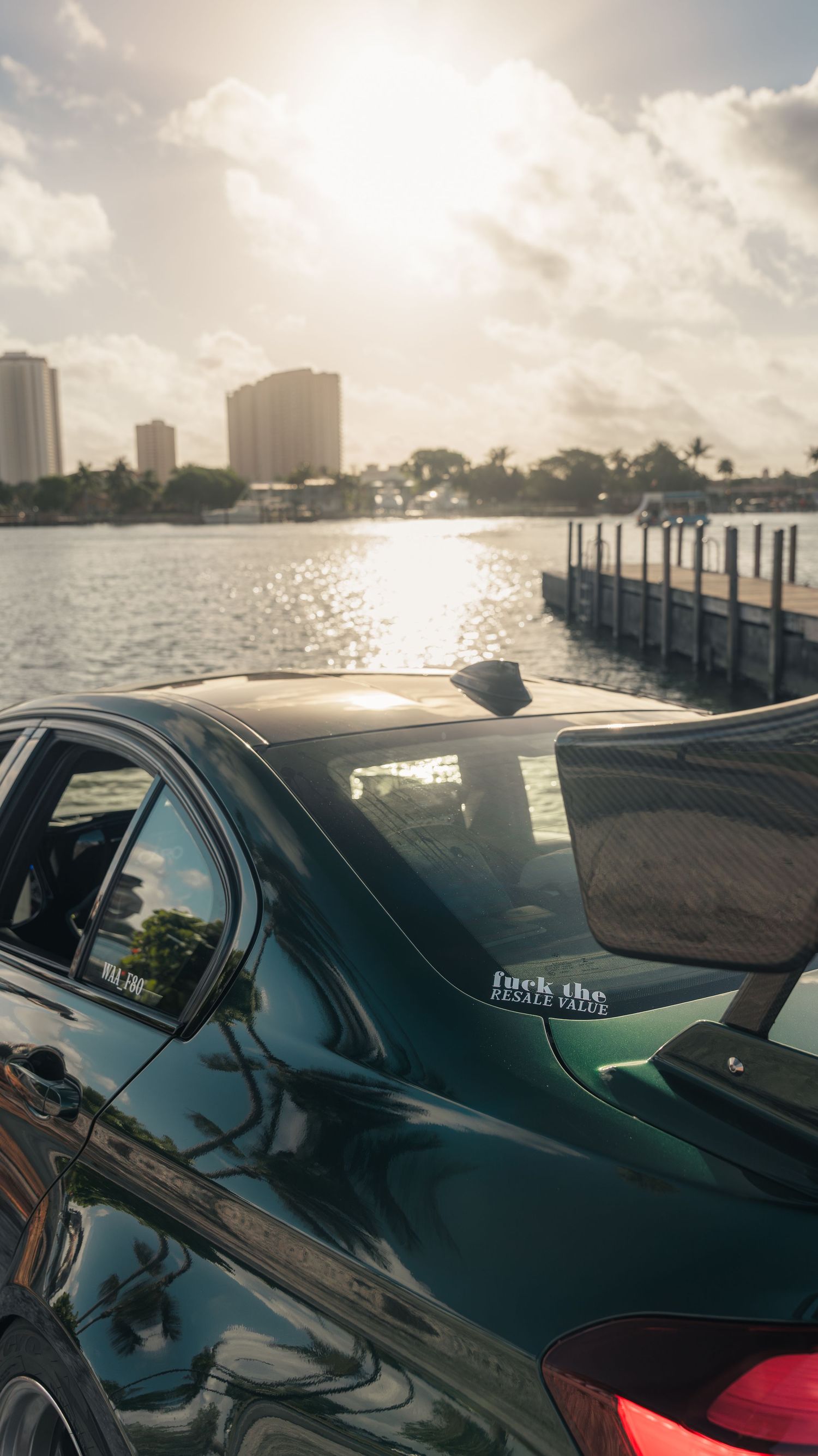 Green car parked near a waterfront pier at sunset with city skyline and reflective water in the background.