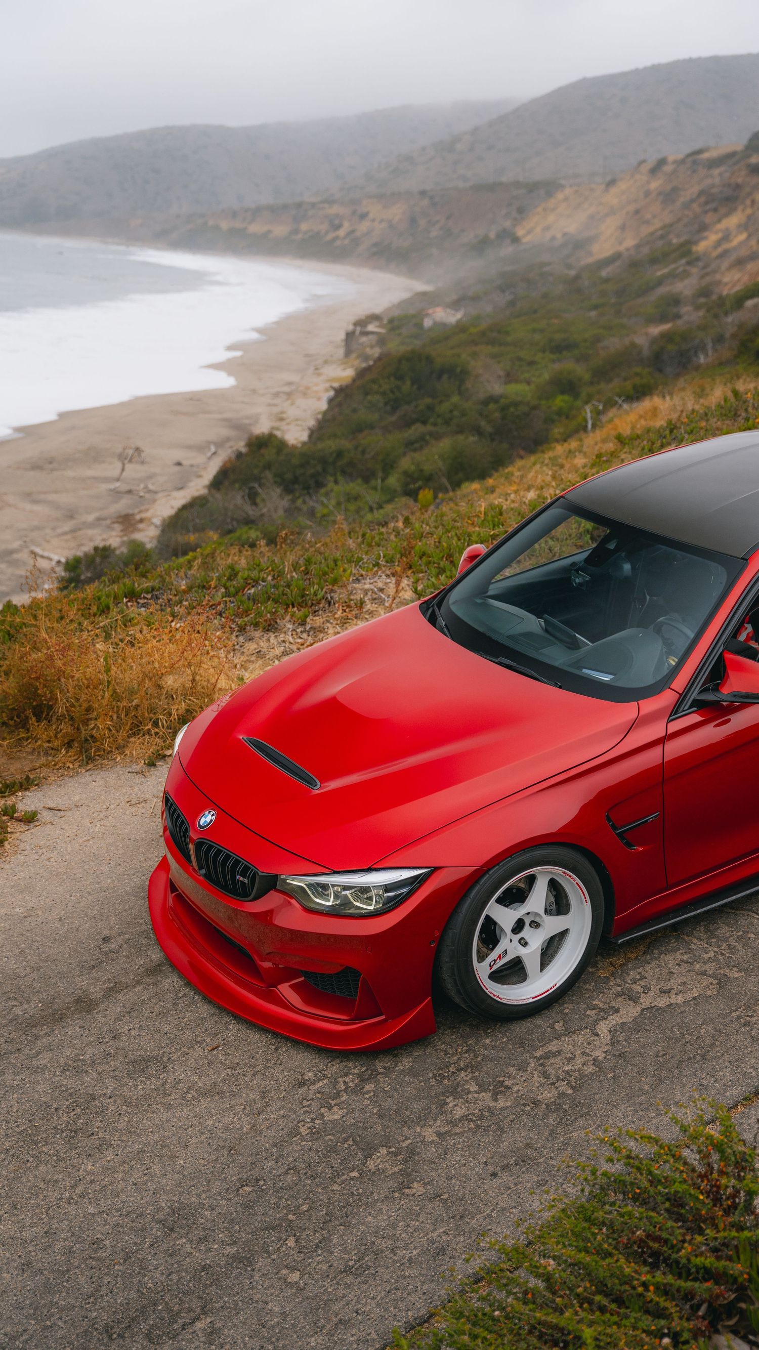 Modified red BMW M4 with white wheels and wide body kit parked on scenic coastal hillside overlook.