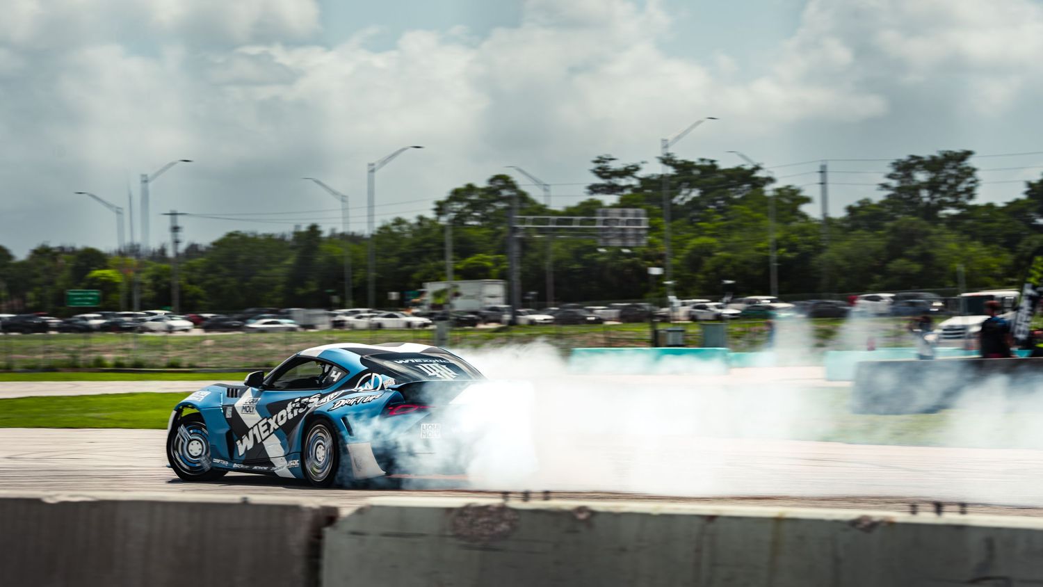 A drift car surrounded by thick tire smoke performs a high-speed drift on a race track with trees and highway in background.