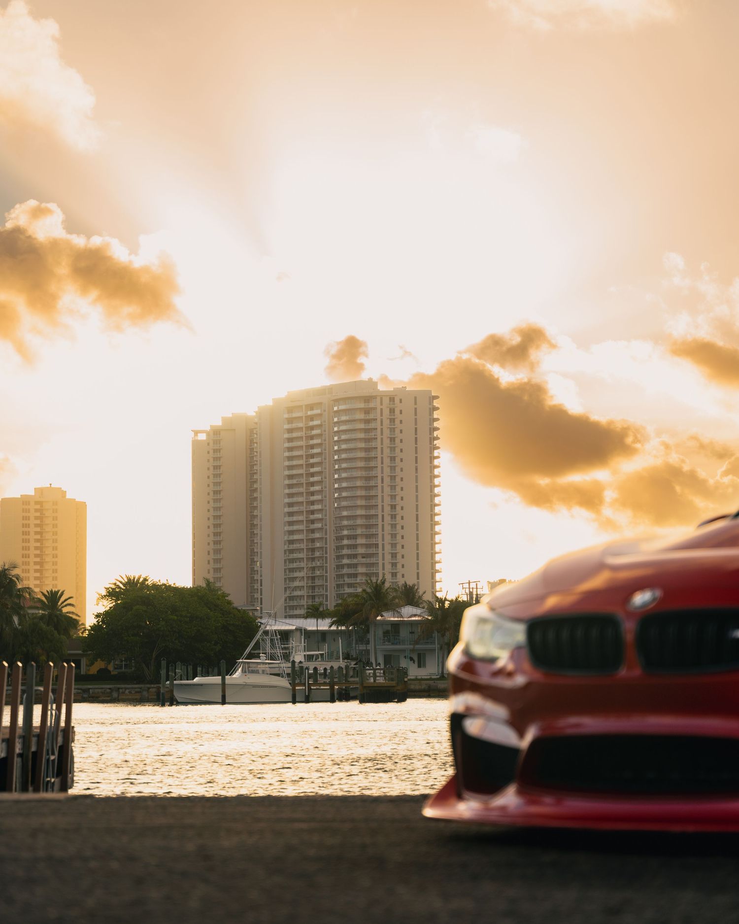 Red BMW parked near a waterfront with luxury high-rise buildings and a golden sunset sky in Miami.