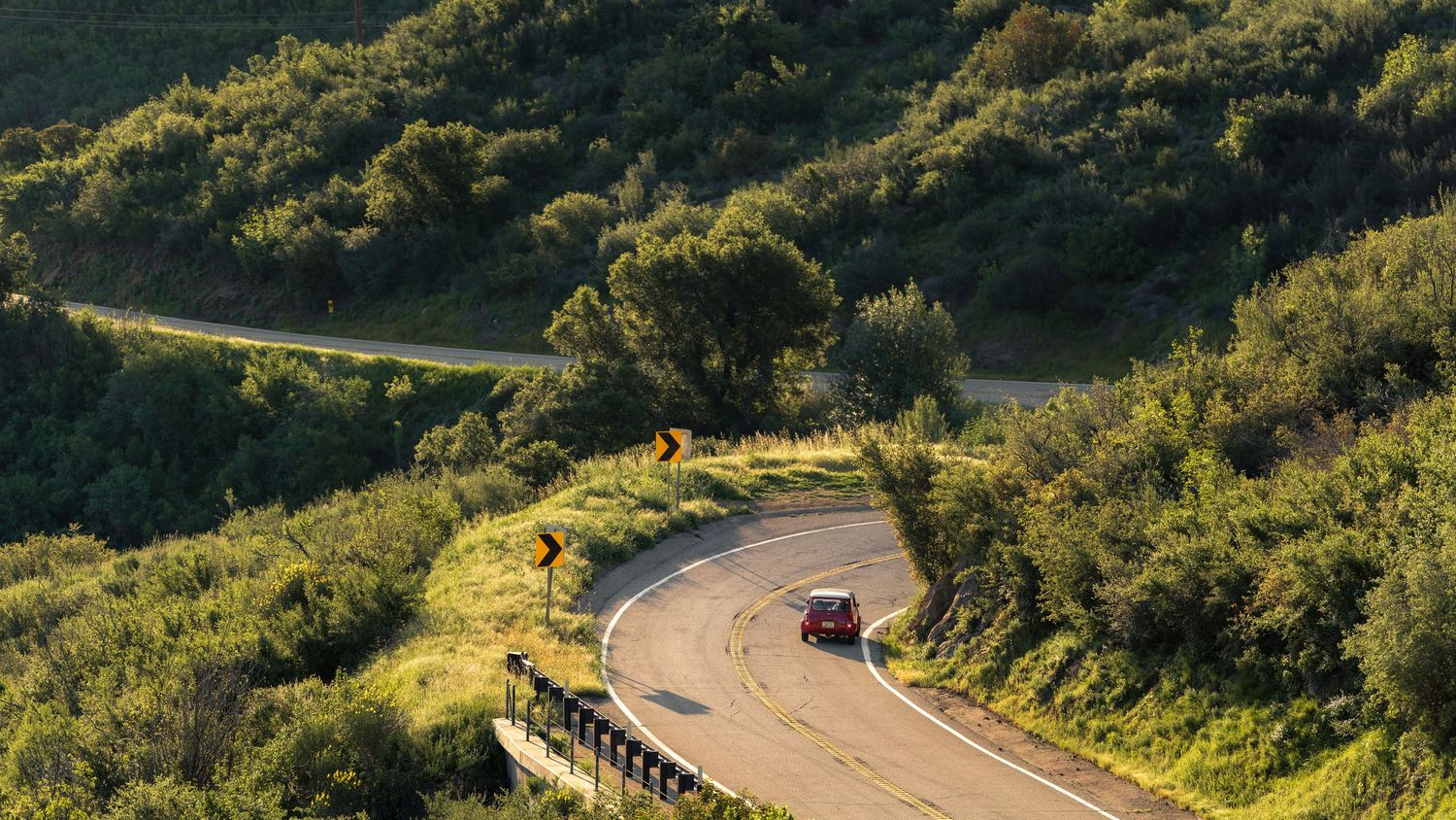 A red classic car navigates a scenic winding mountain road with rocky cliffs and lush vegetation.