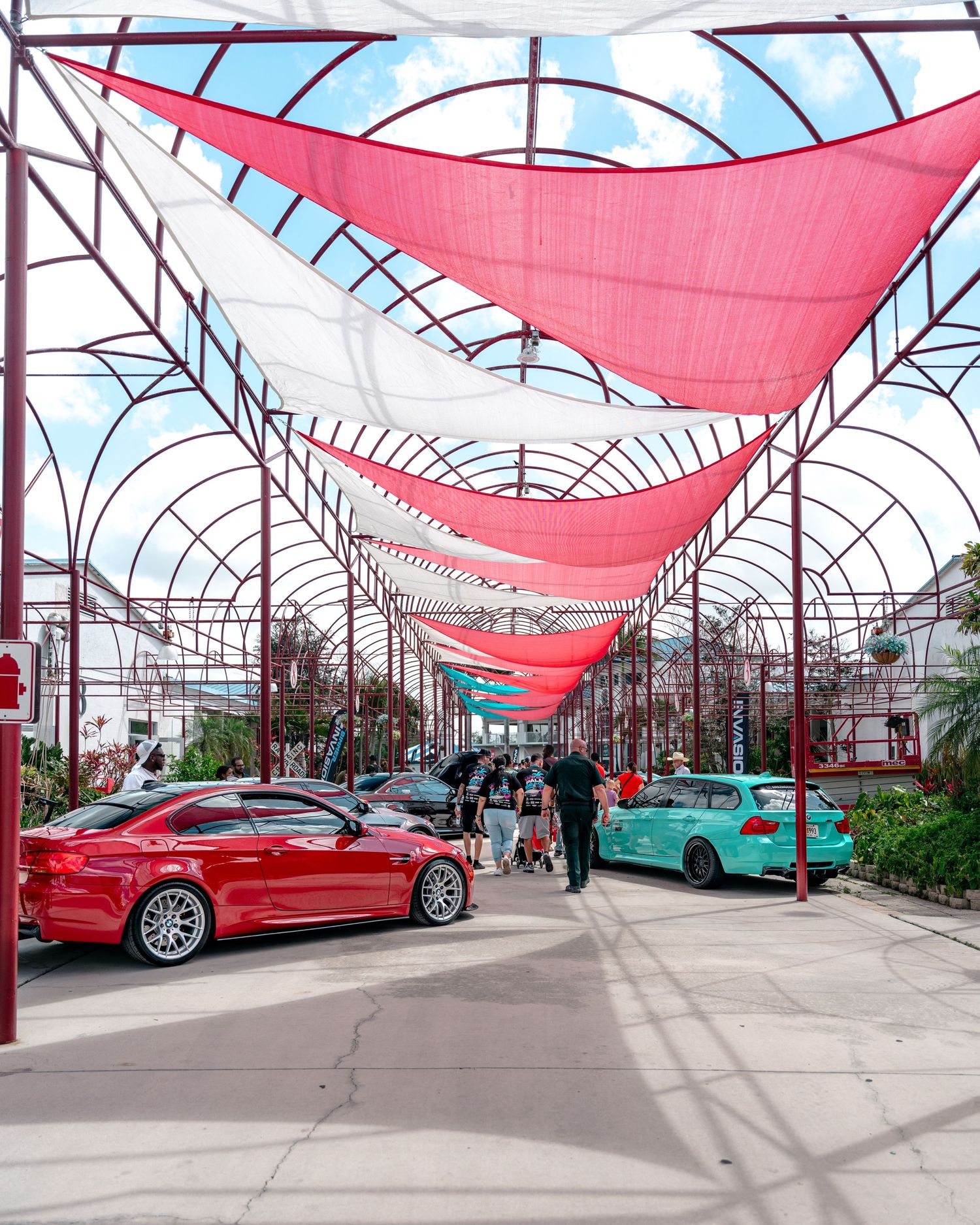 Colorful red and white shade sails stretch over a car show corridor with a red BMW and teal wagon on display.