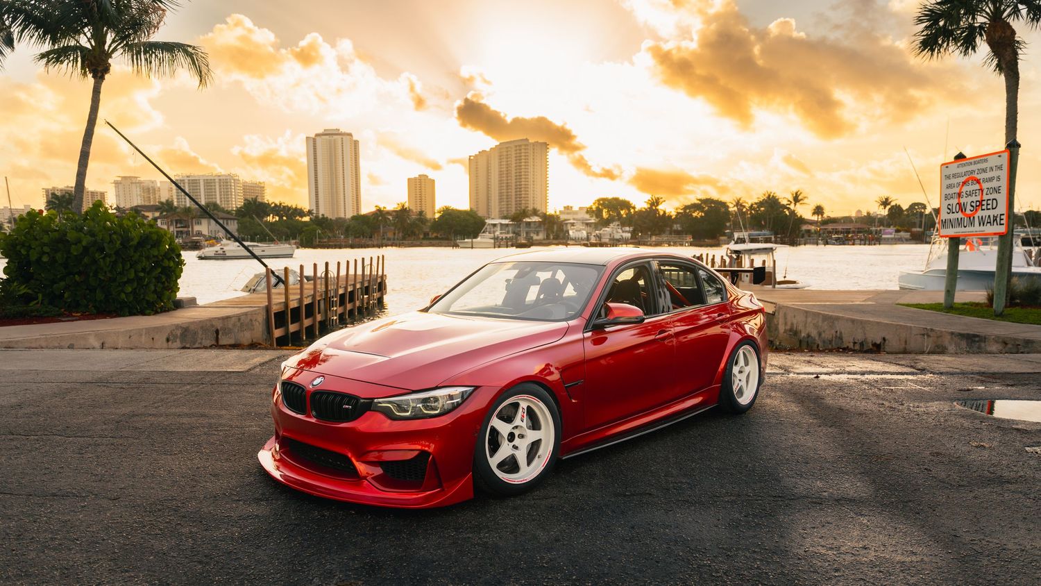 Red BMW M3 on white wheels parked near waterfront at sunset with city skyline and palm trees in background.