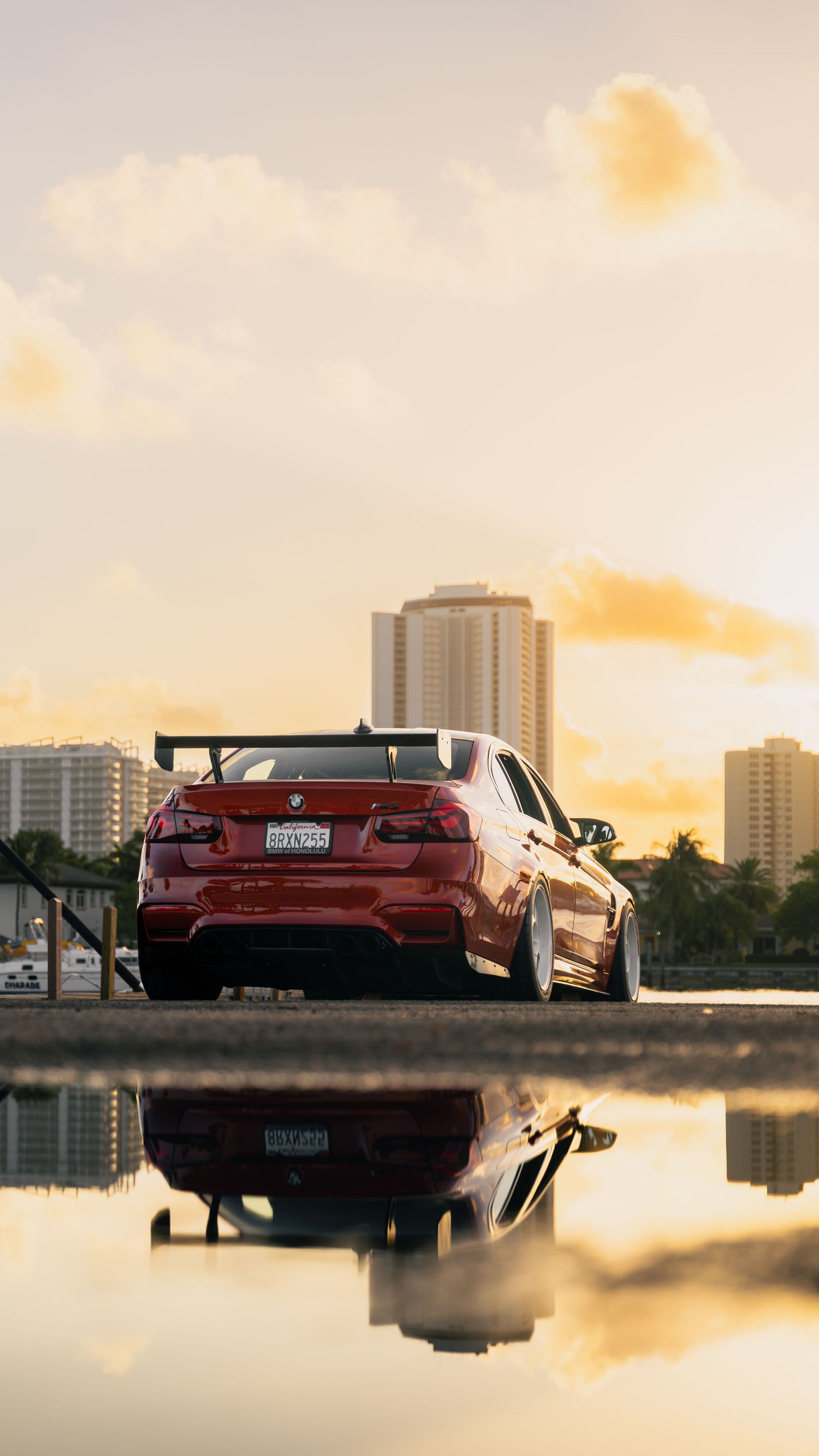 Red BMW M3 with rear spoiler reflected in water at golden hour with city skyline backdrop.