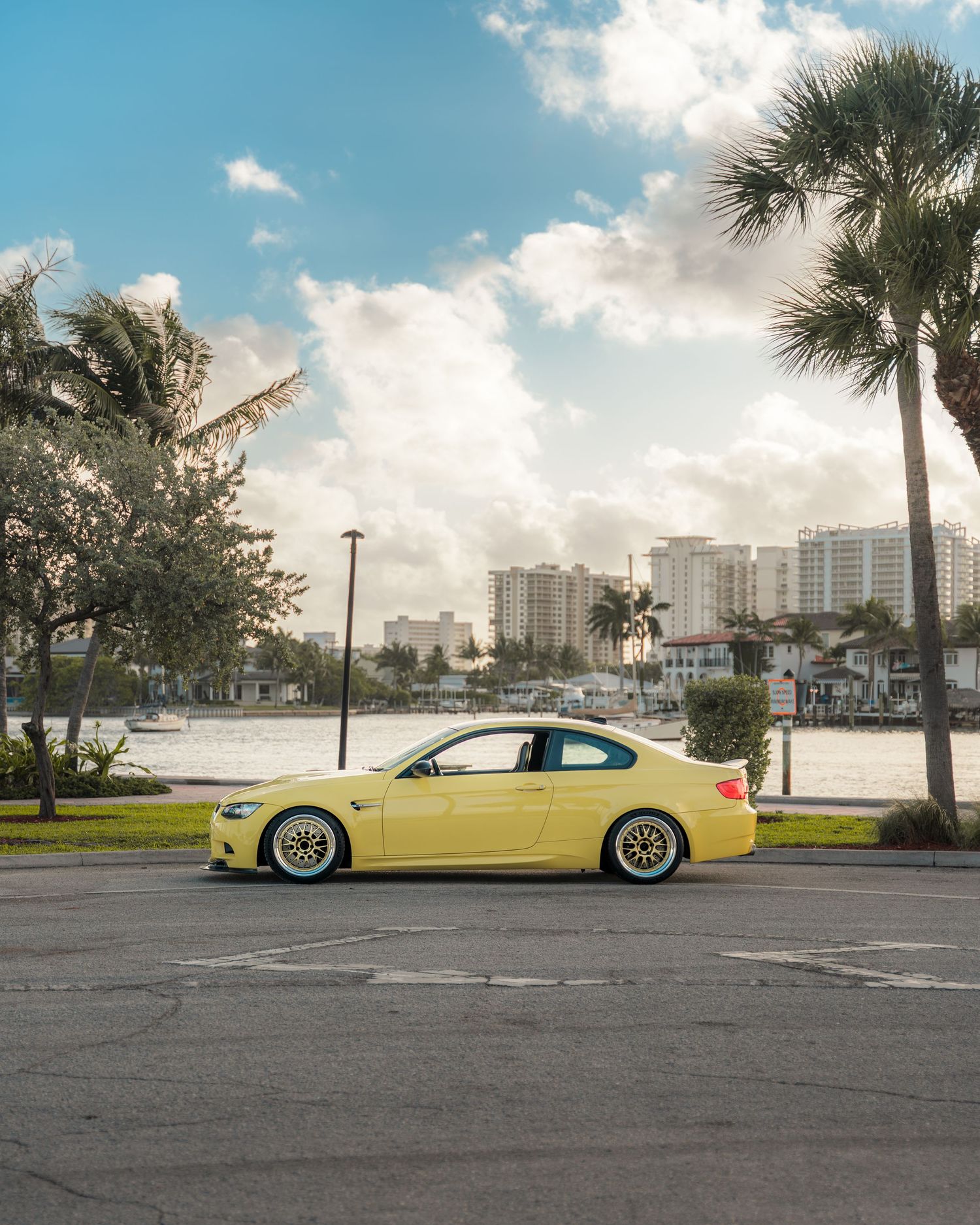 Yellow BMW coupe parked near waterfront with palm trees and city skyline under partly cloudy sky in Florida.