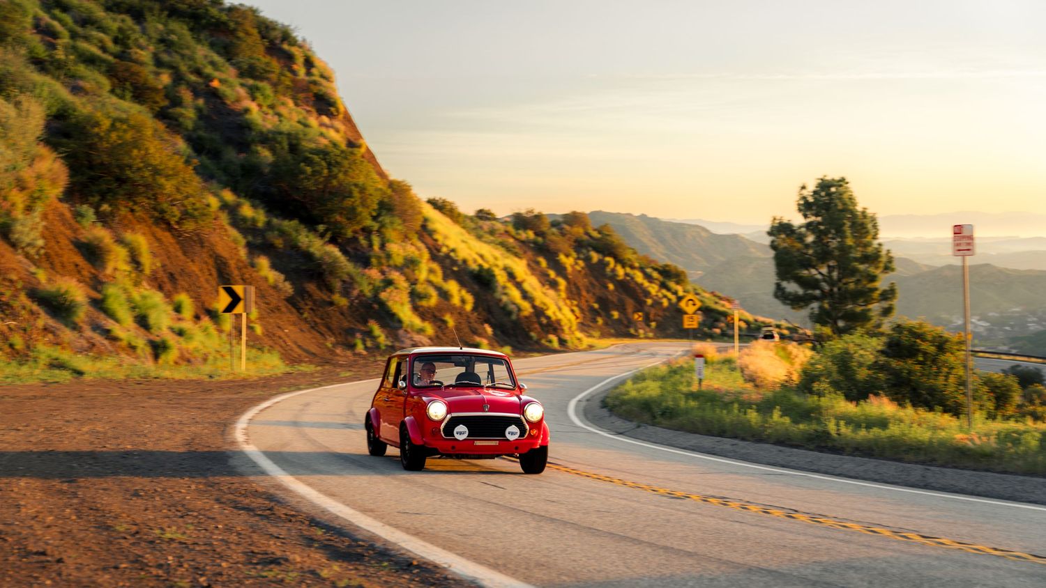 Classic sports car navigates a winding mountain road during golden hour sunset with scenic coastal views.