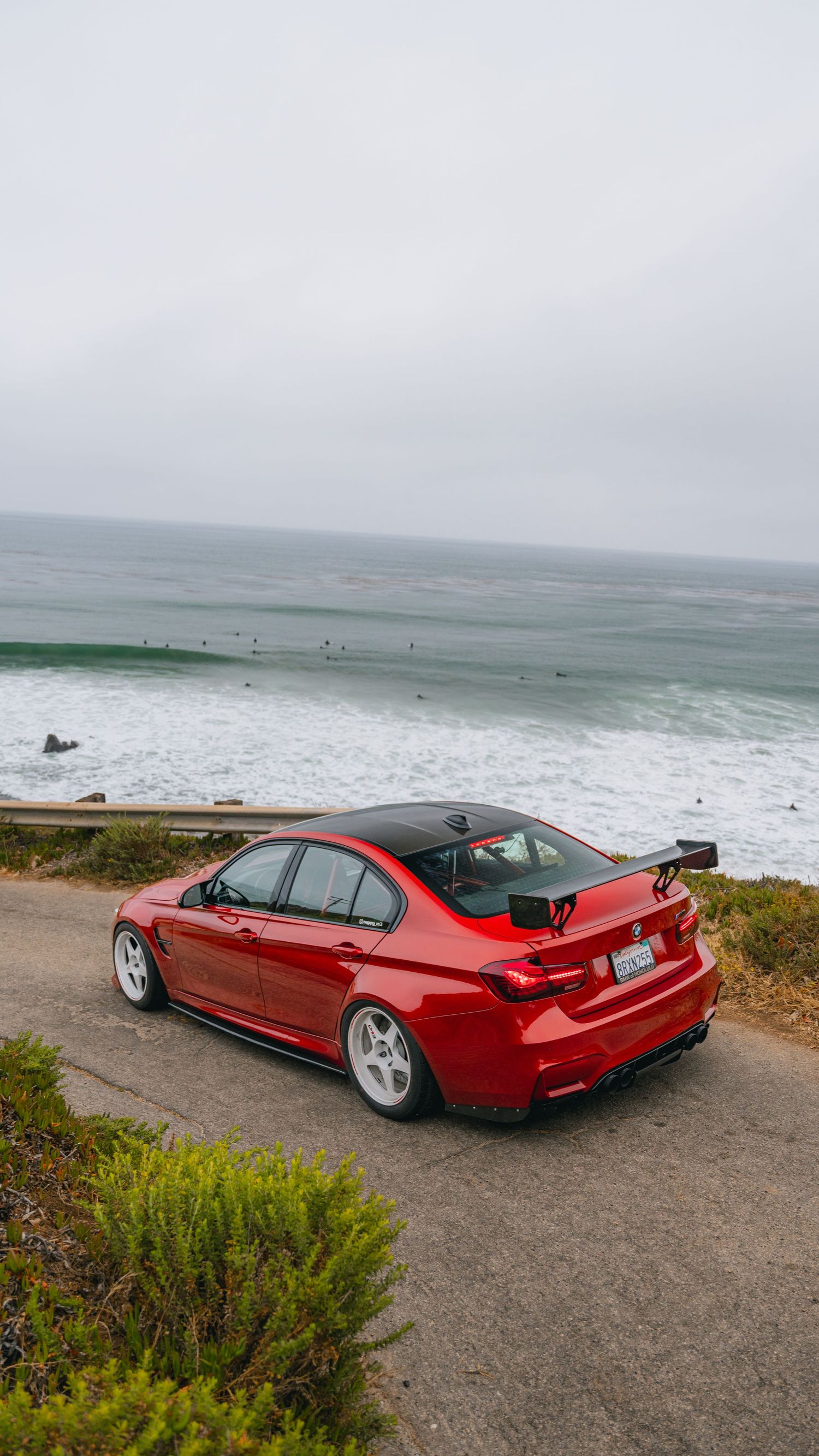 Red BMW M3 with rear spoiler parked on scenic overlook above ocean coastline.