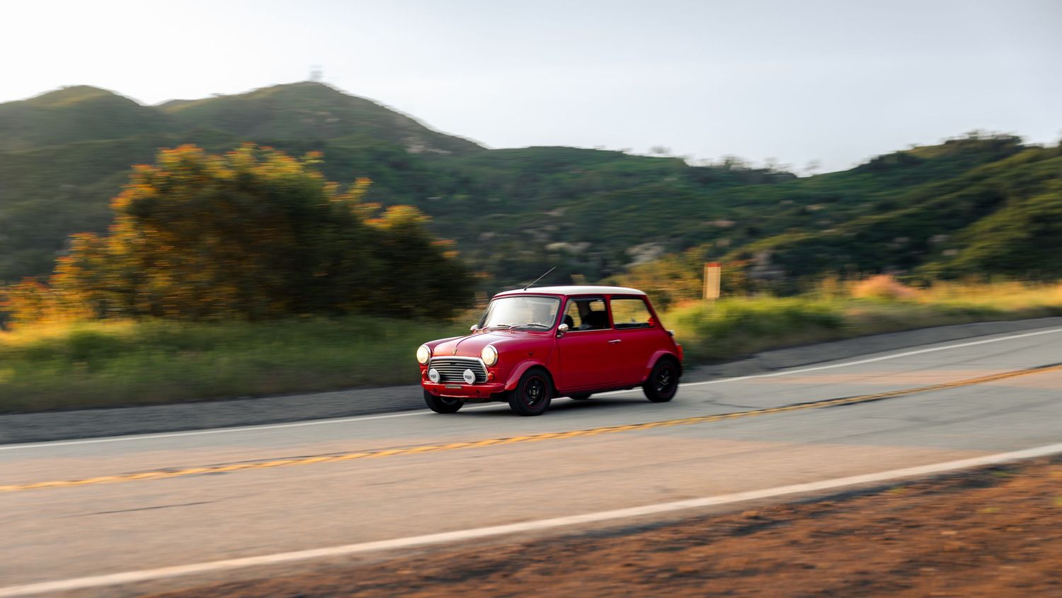 A red vintage car drives along a winding mountain road with scenic views of hills and valleys in the background.