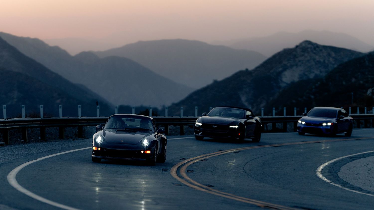 Panoramic view of sports cars cruising on a mountain highway against a dusky skyline.