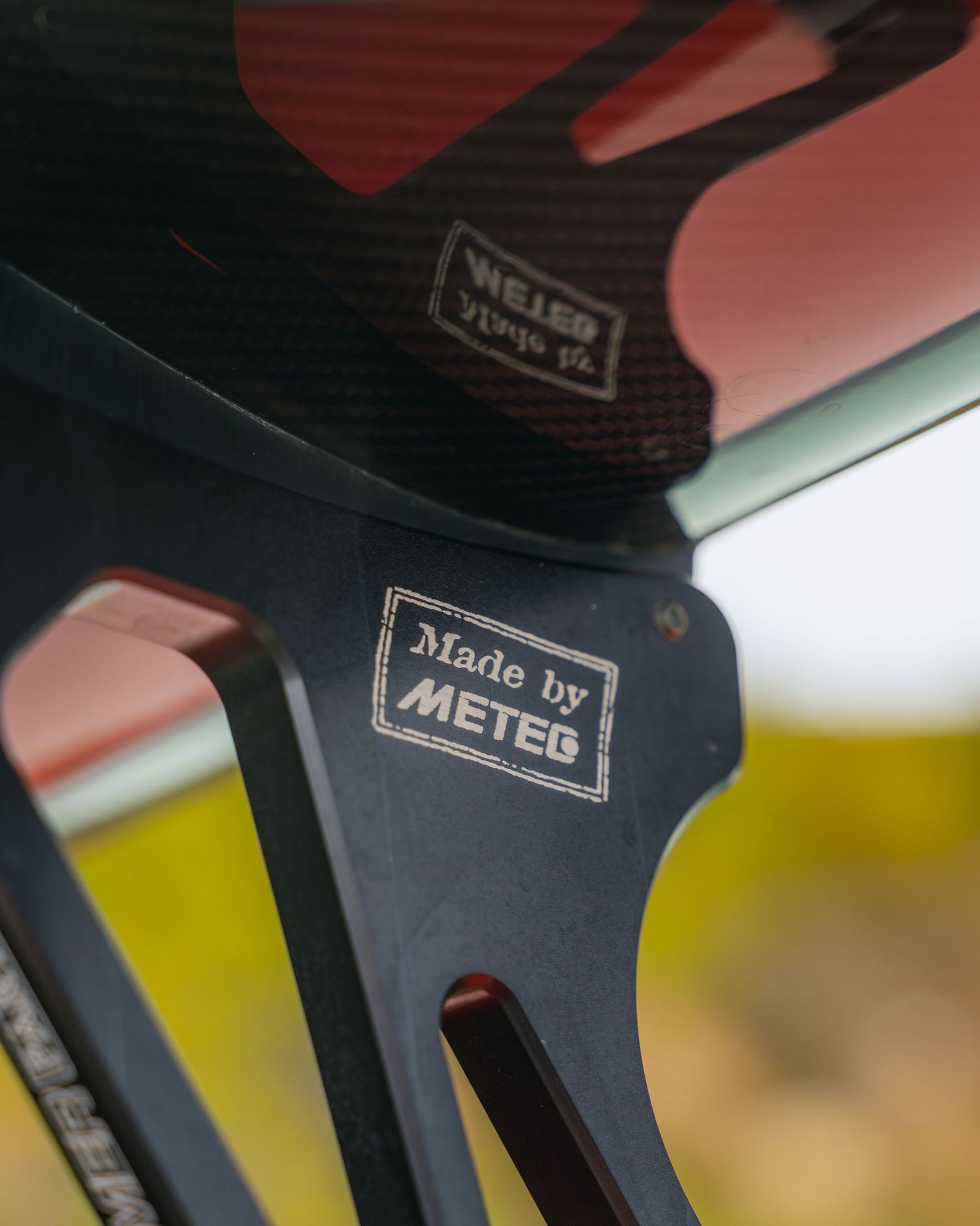 Close-up of a black bicycle component with an engraved Made by METEG label, featuring carbon fiber details in the background.