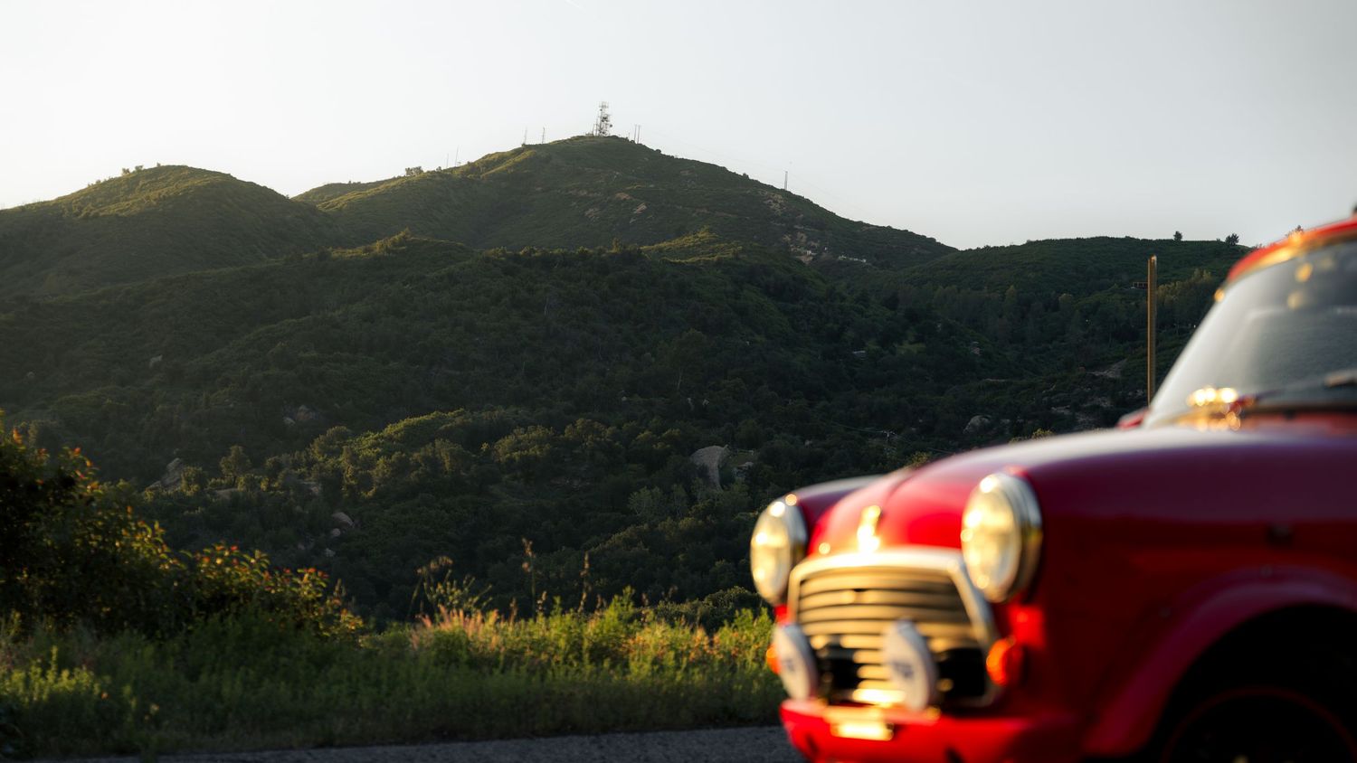 Red vintage car parked on hillside during sunset with mountains in the background.