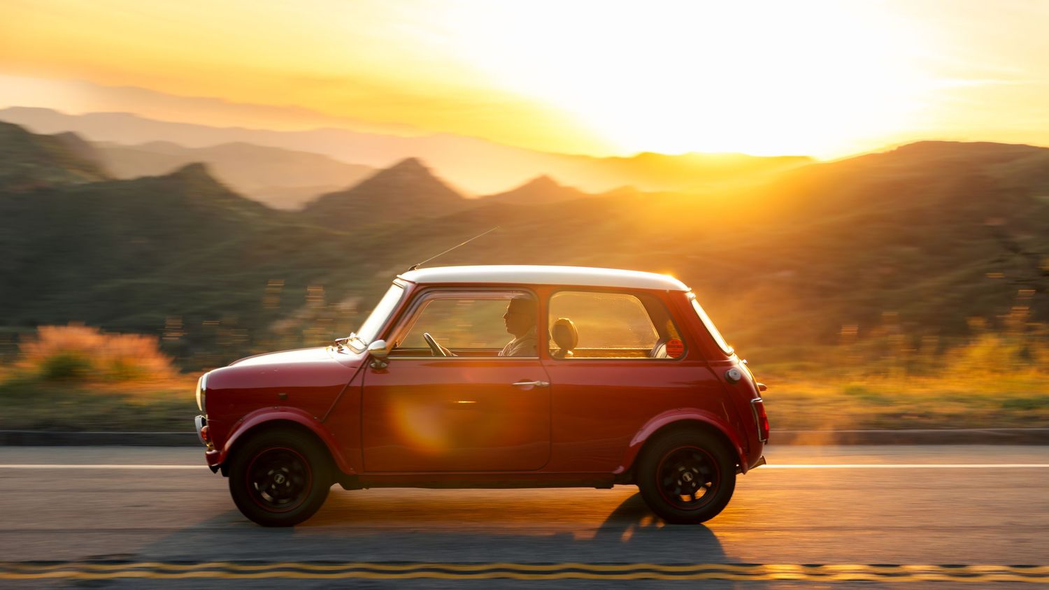 A classic Mini Cooper car silhouetted against a dramatic mountain sunset with golden light illuminating the scene.