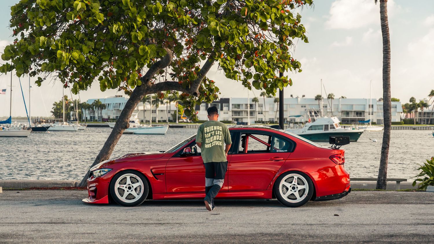 Man standing beside a lowered red BMW 3 Series near waterfront with trees and boats in background.