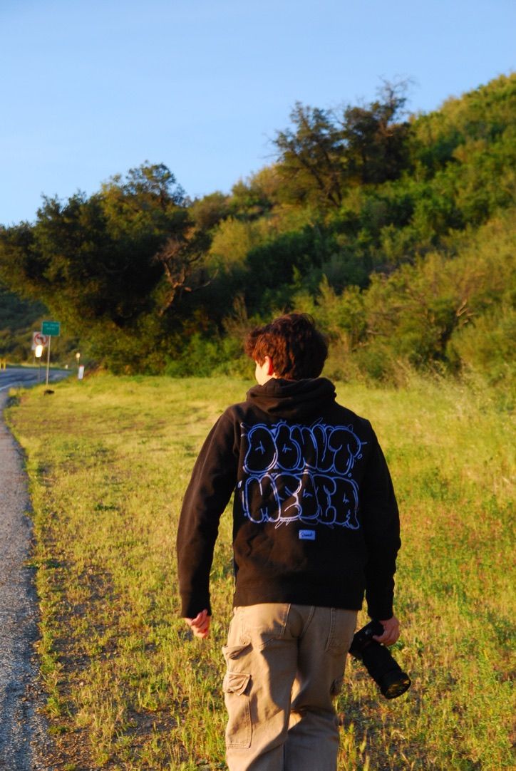Person walking along a rural road wearing a black graphic hoodie, carrying a camera, with green hills in the background.