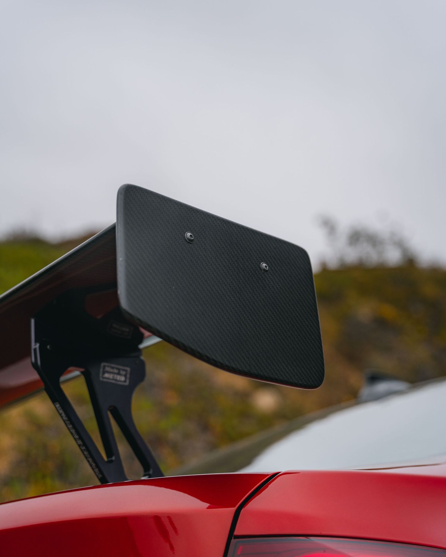 Carbon fiber rear spoiler wing on a red BMW performance car against a cloudy sky backdrop.