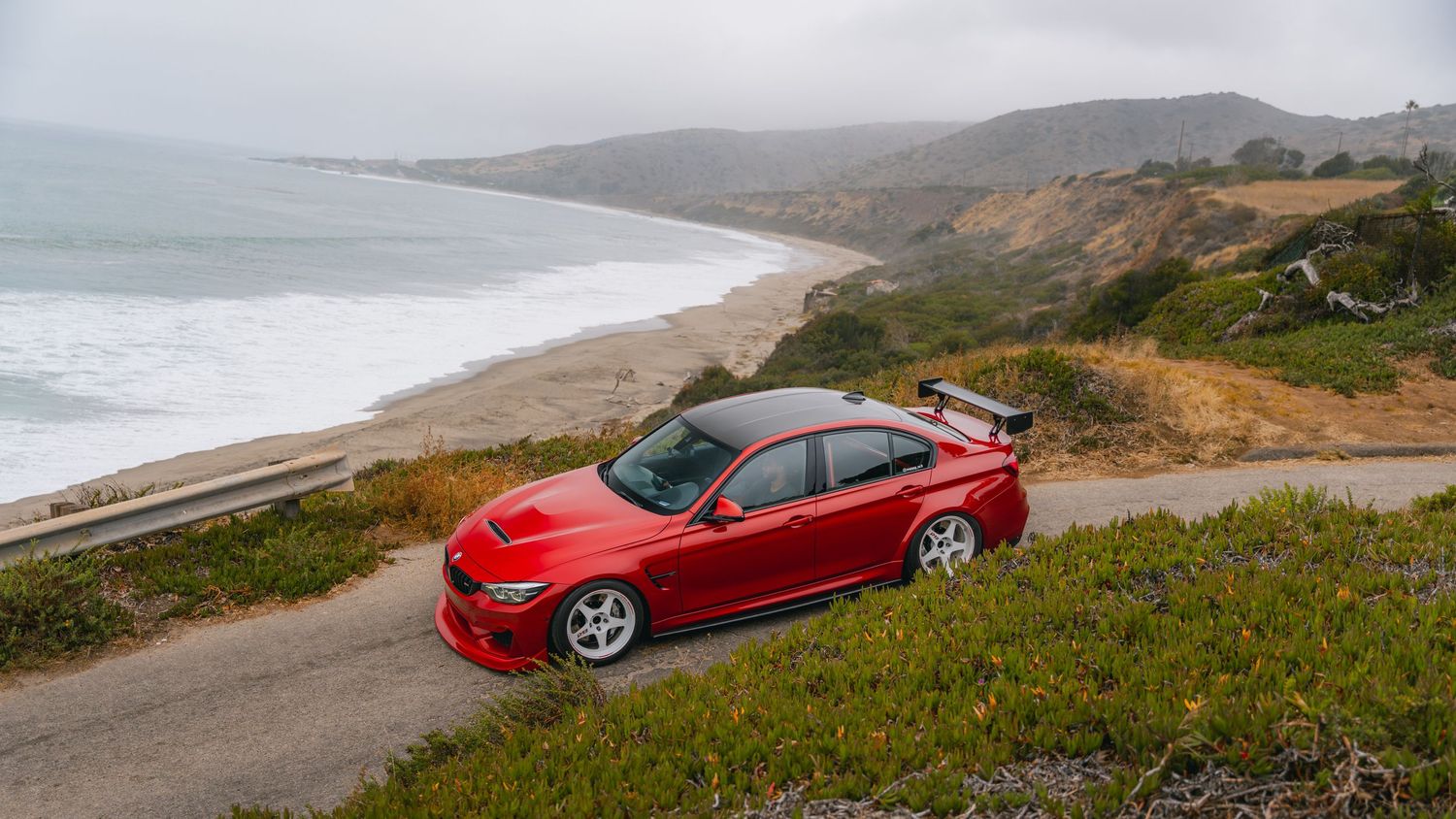 Red Mitsubishi Lancer Evolution parked on a coastal road with ocean views and misty hills in the background.