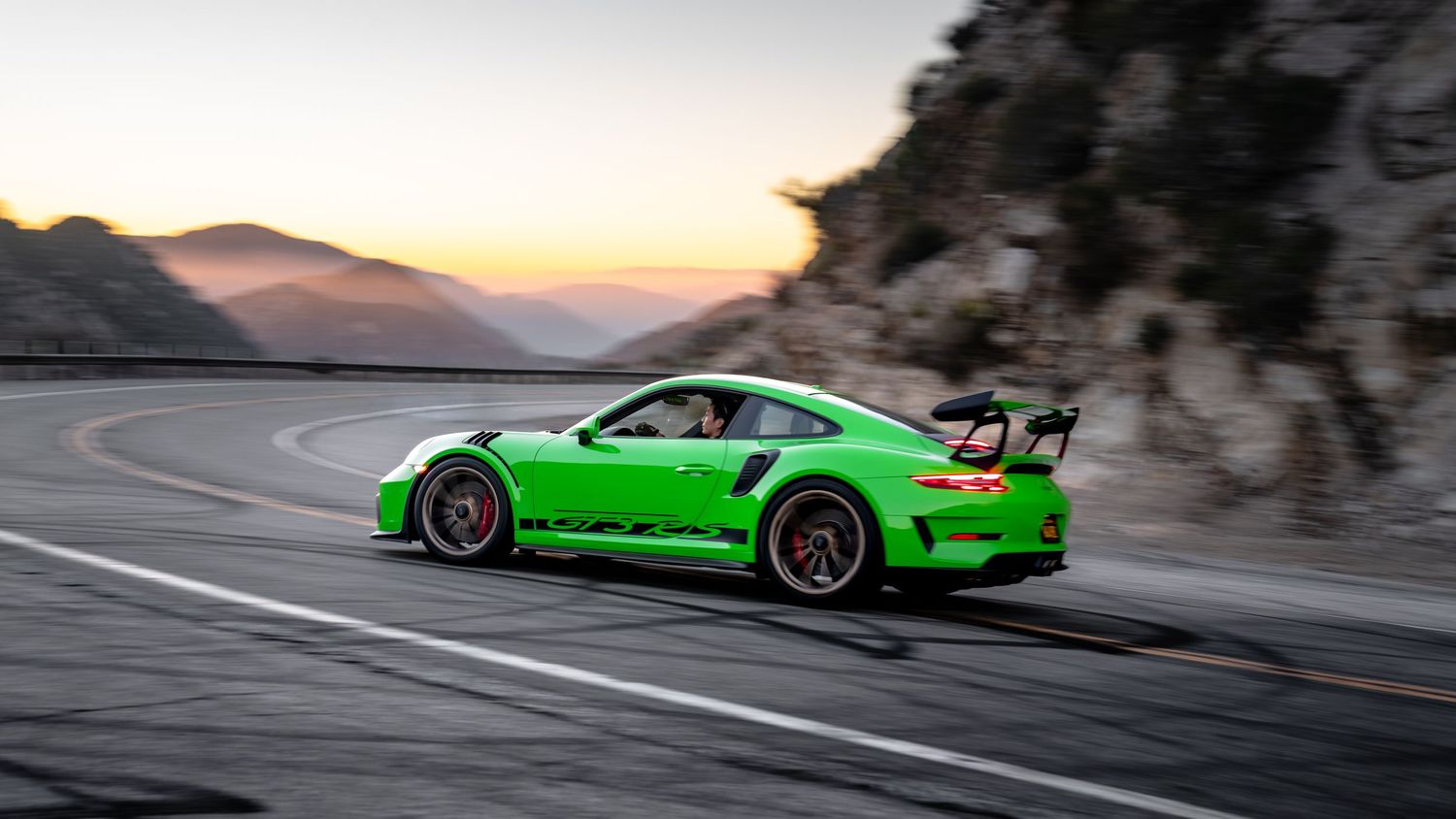 Bright green sports car driving along a curving mountain road at sunset.