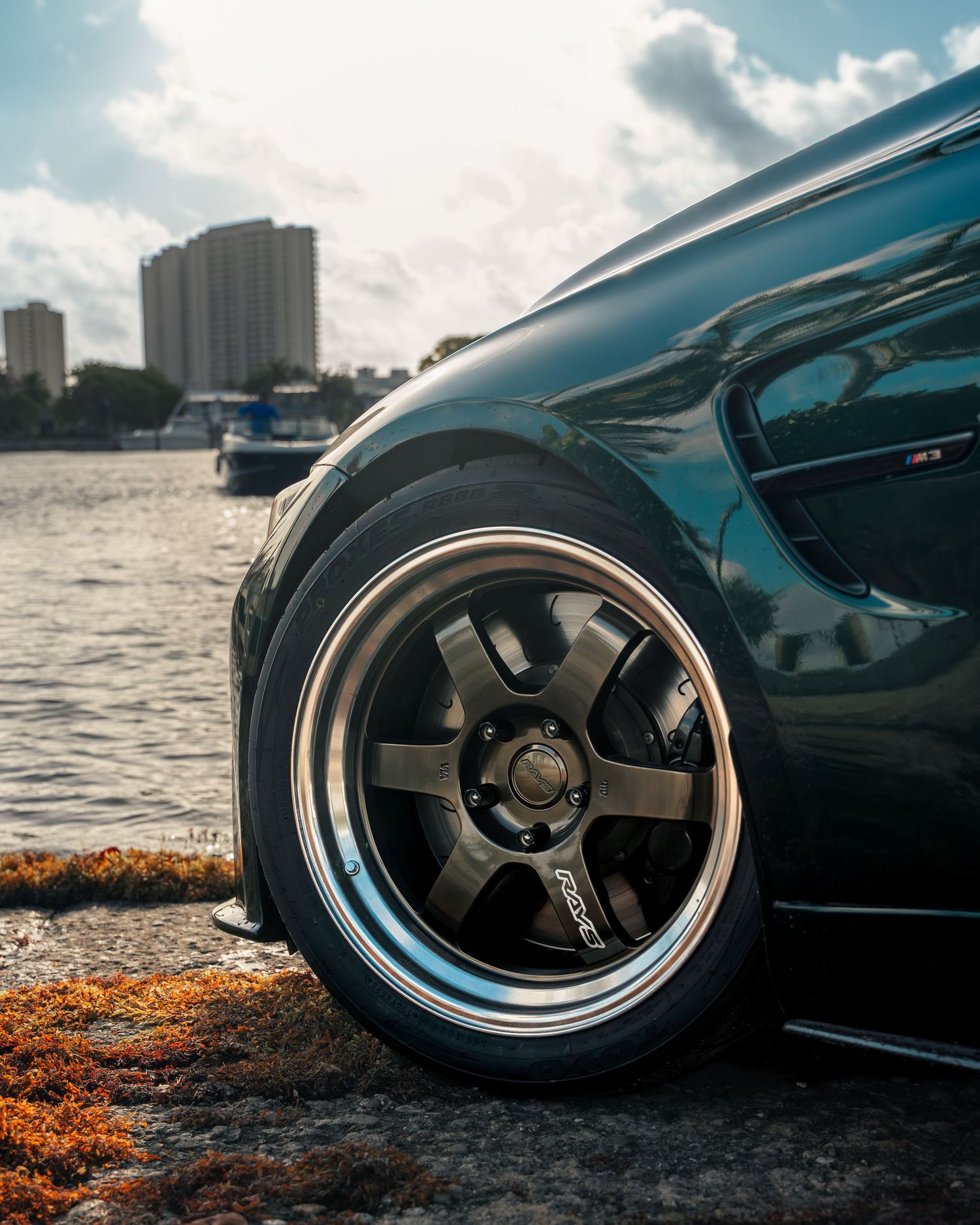 Dark green sports car with bronze multi-spoke aftermarket wheels parked near waterfront on sunny day.