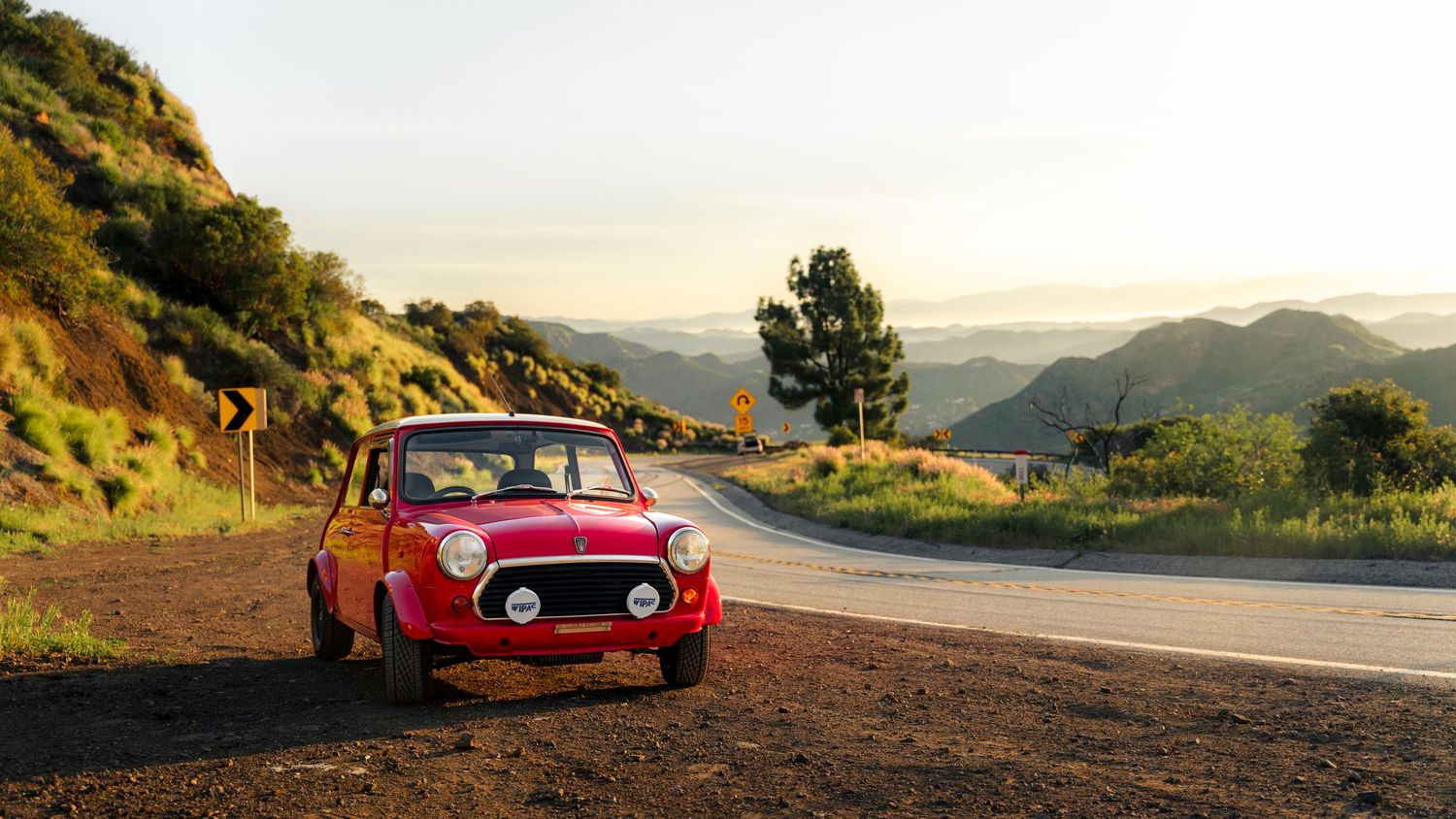 A vintage red sports car parked on a scenic mountain road at sunset with rolling hills in the background.