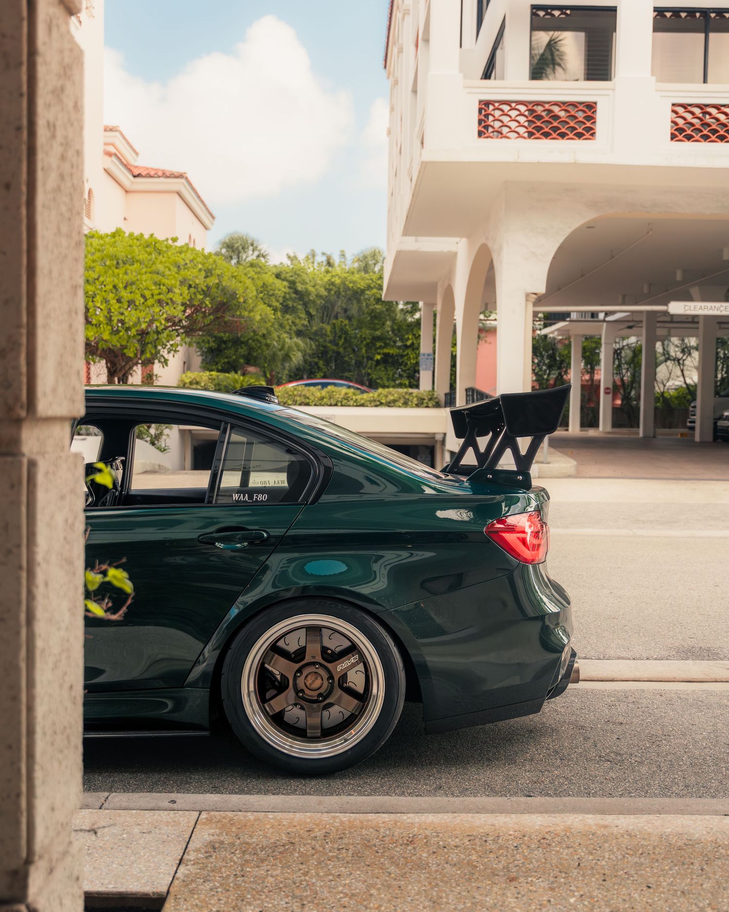 Dark green modified sedan with large rear wing and custom wheels parked near a white building on a sunny day.