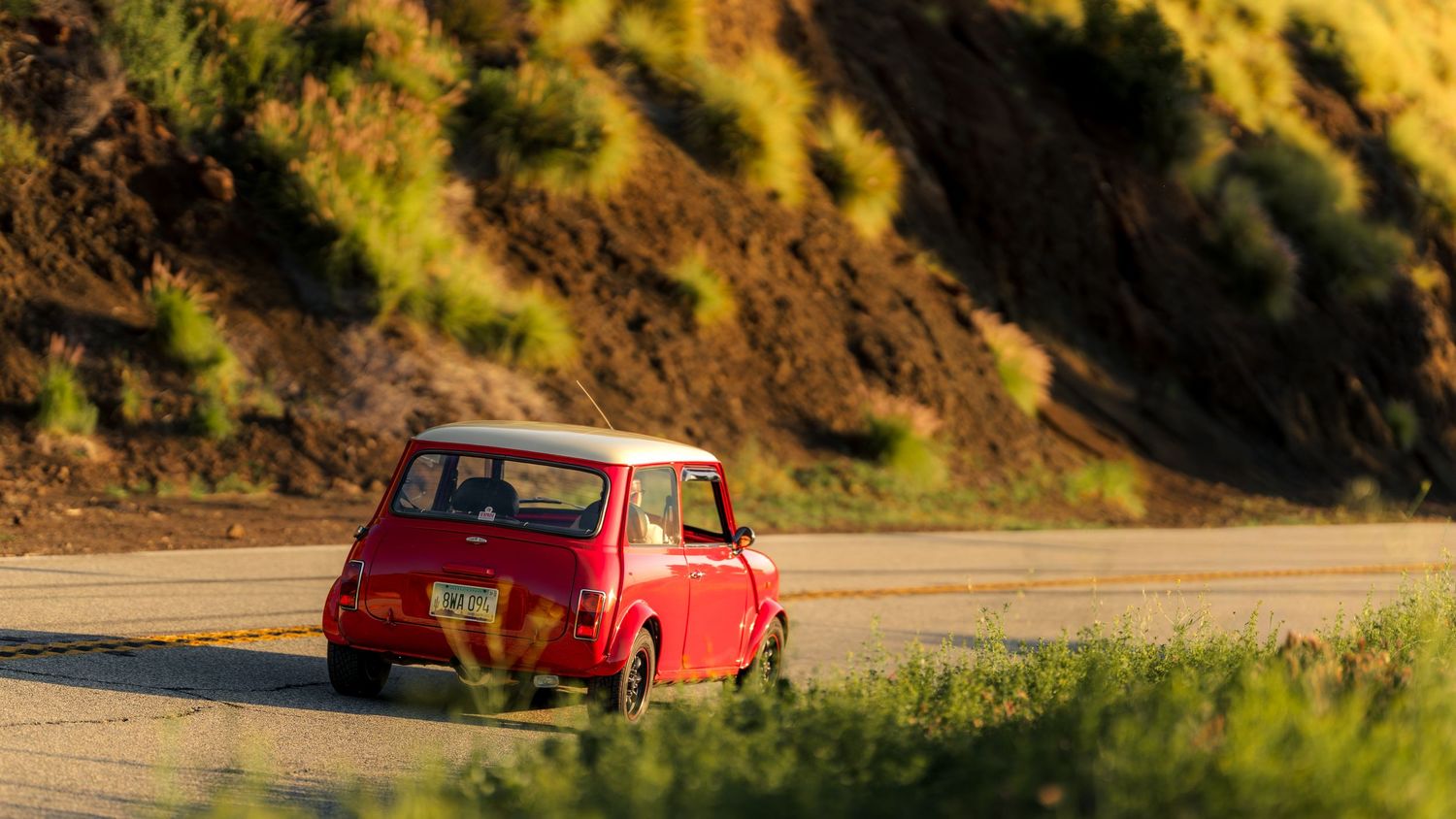 A red vintage car drives along a scenic mountain road with golden autumn foliage in the background.