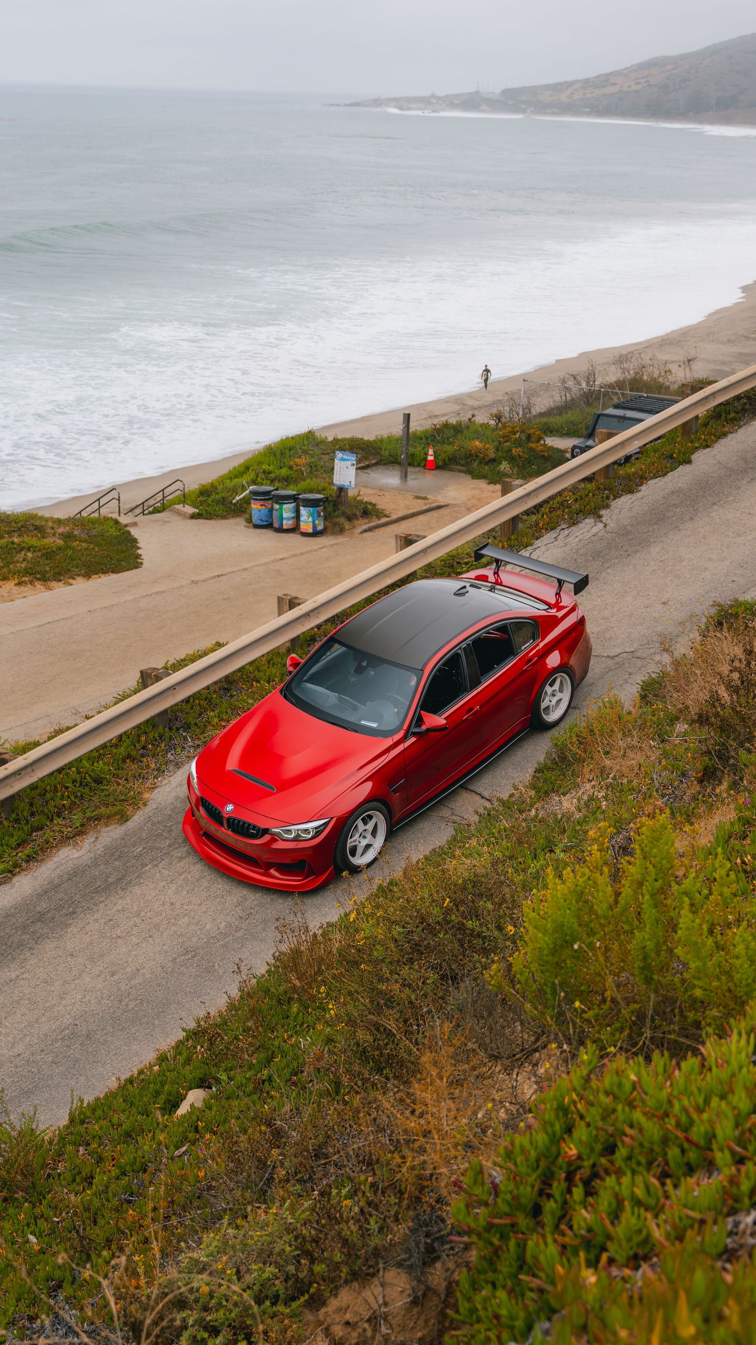 Red BMW M4 with carbon roof and rear spoiler parked on coastal road overlooking sandy beach and ocean.