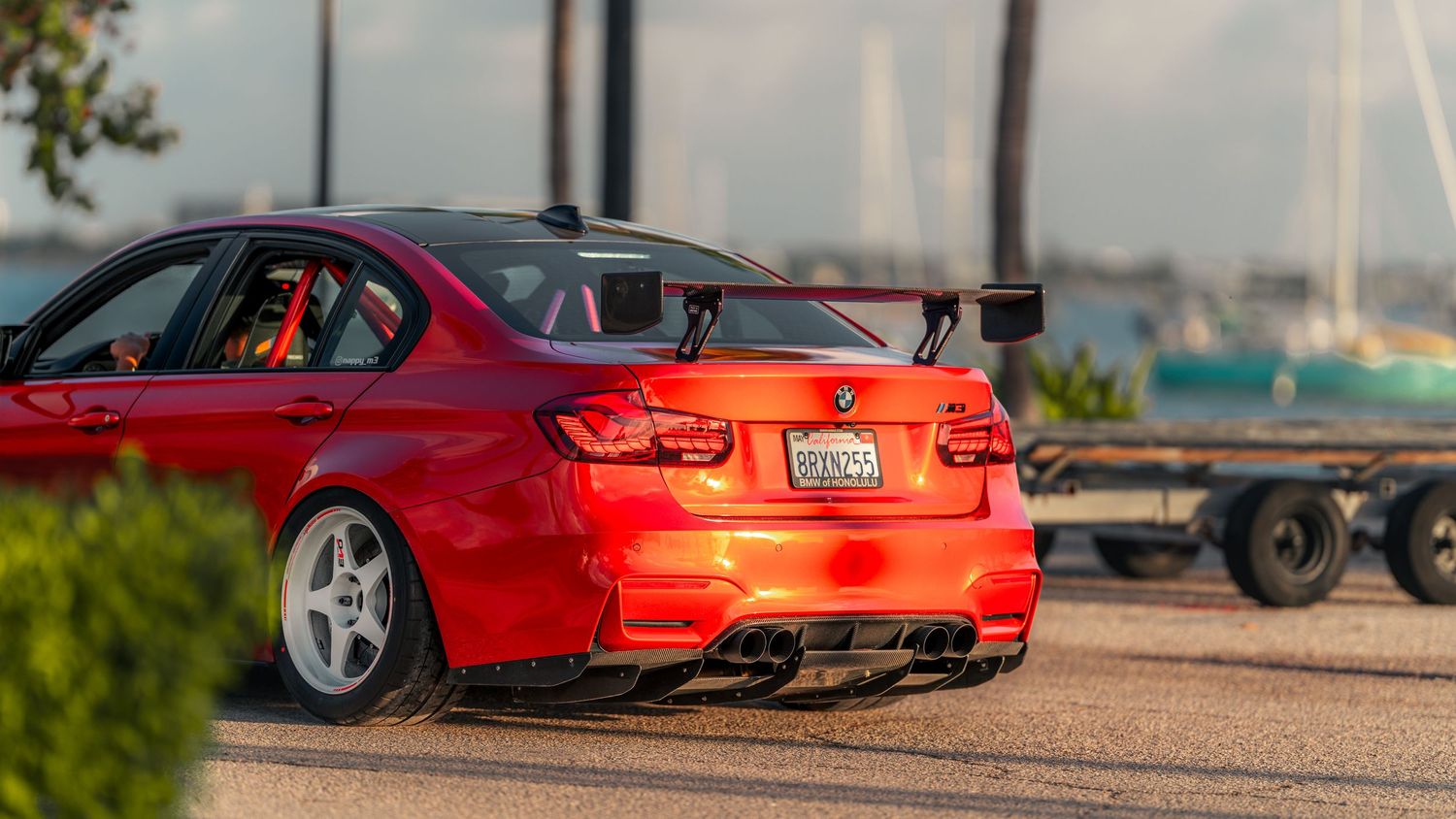 Red BMW M3 sedan with carbon fiber spoiler and diffuser parked near waterfront on a sunny day.